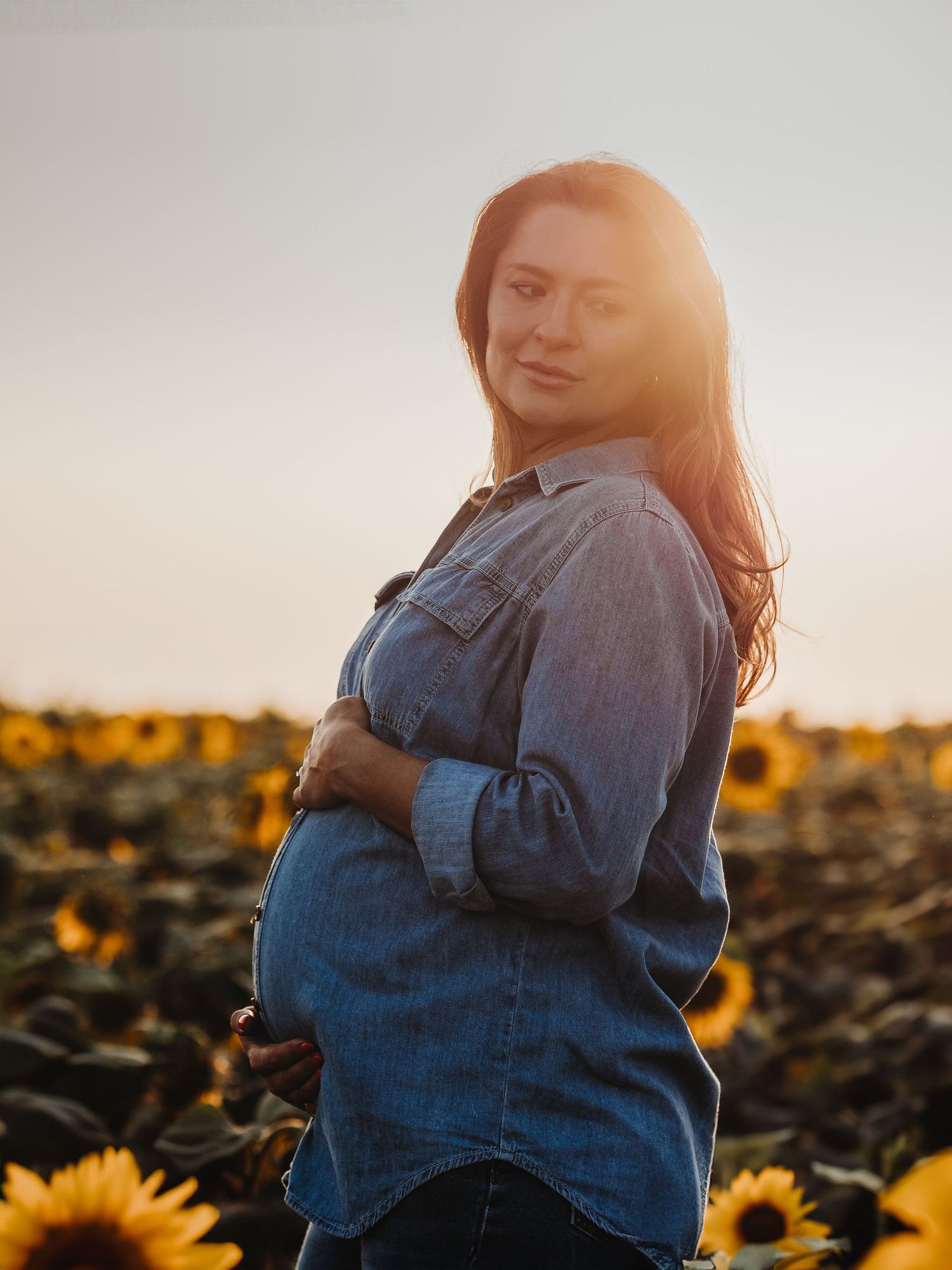 A figure stands in a sunflower field at golden hour, wearing a denim shirt with sunlight creating a warm backlit glow.
