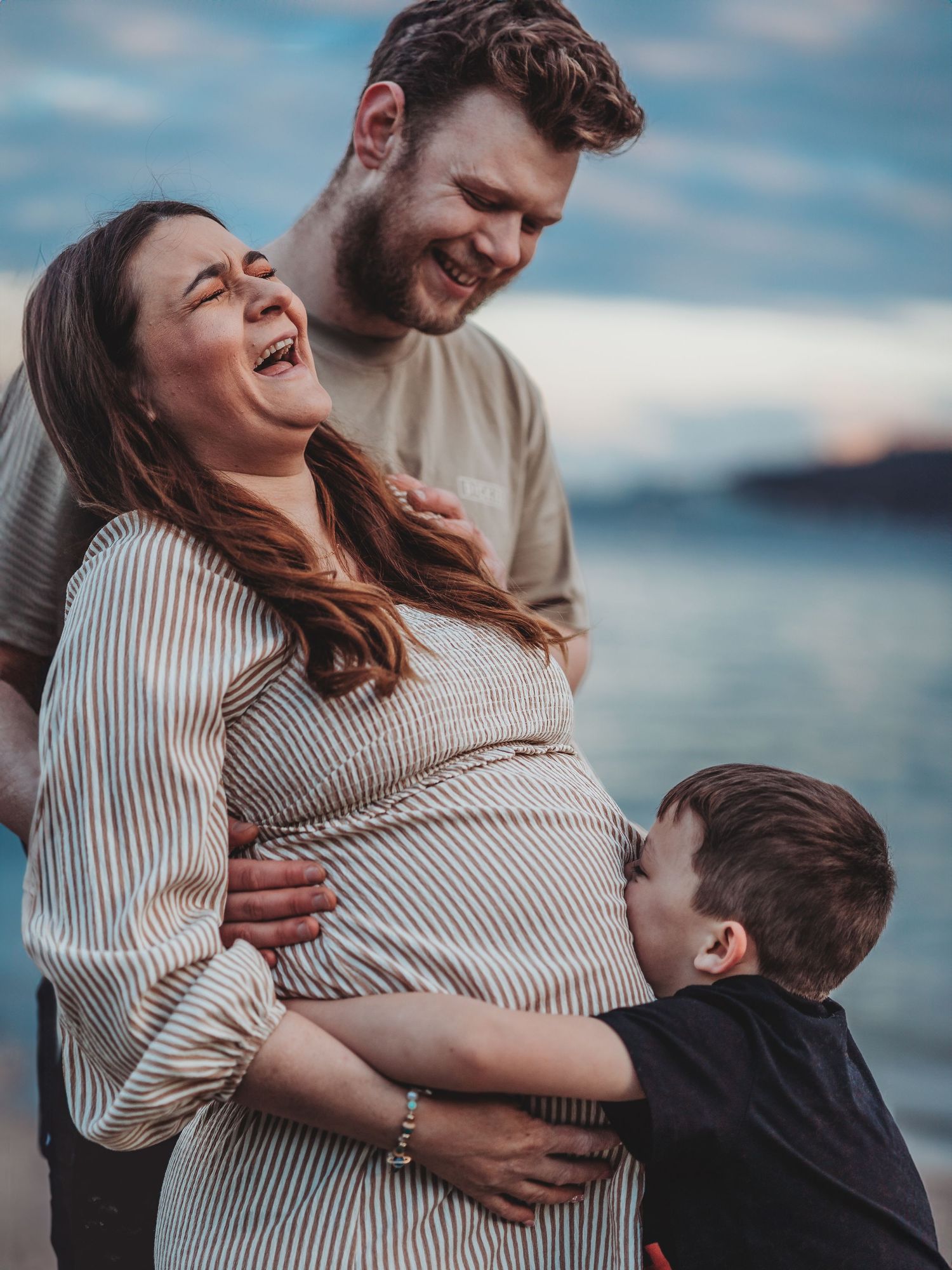 A couple and child share joyful moments by the water during a maternity photoshoot at sunset.
