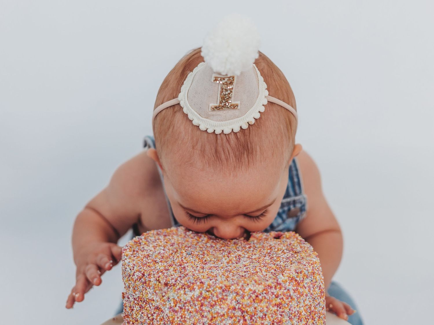 Baby wearing a white bib smashes and plays with a pink frosted birthday cake during a cake smash photo session.