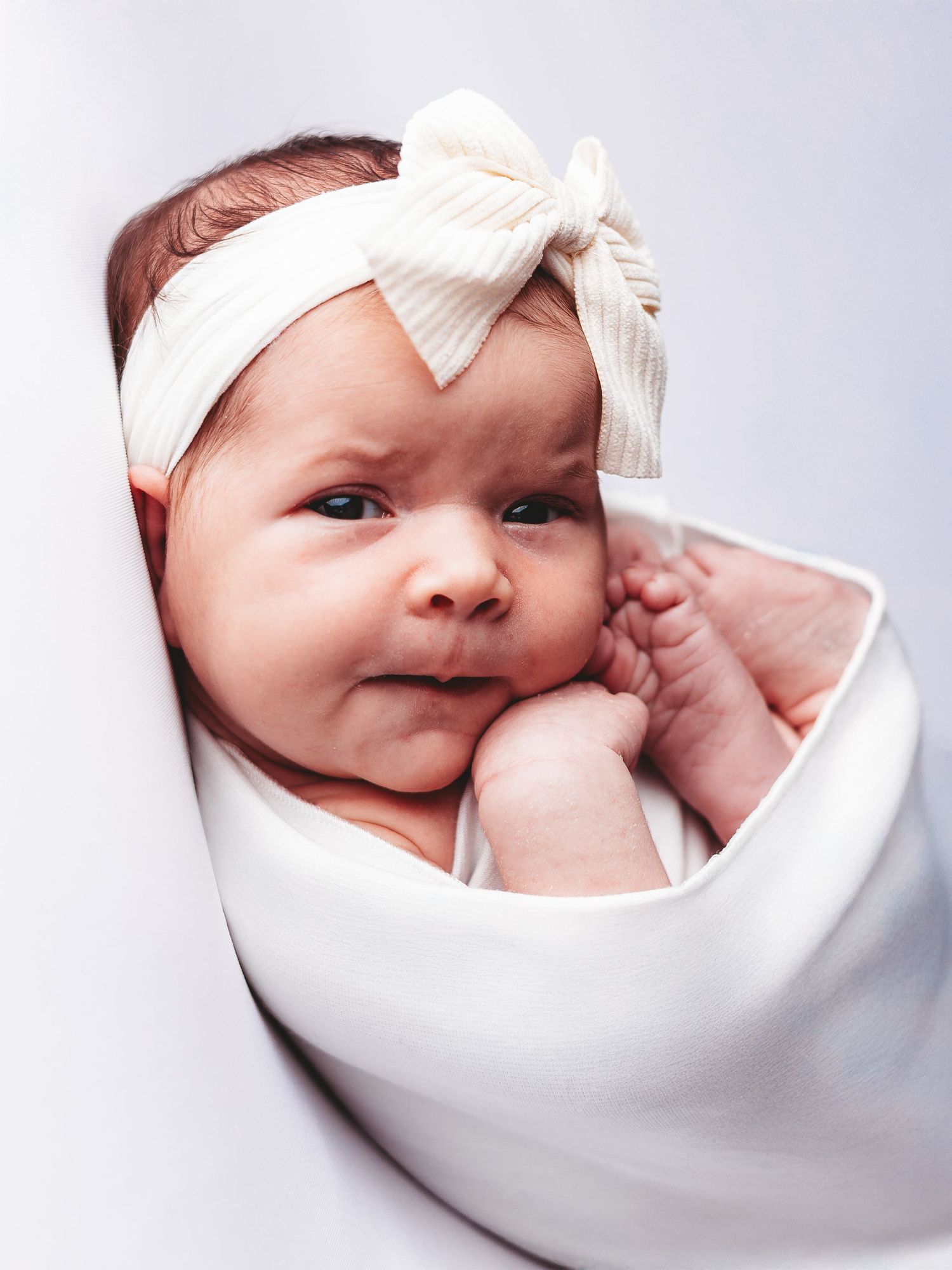 A baby wrapped in a white swaddle blanket wearing a white bow headband makes an adorable expression.