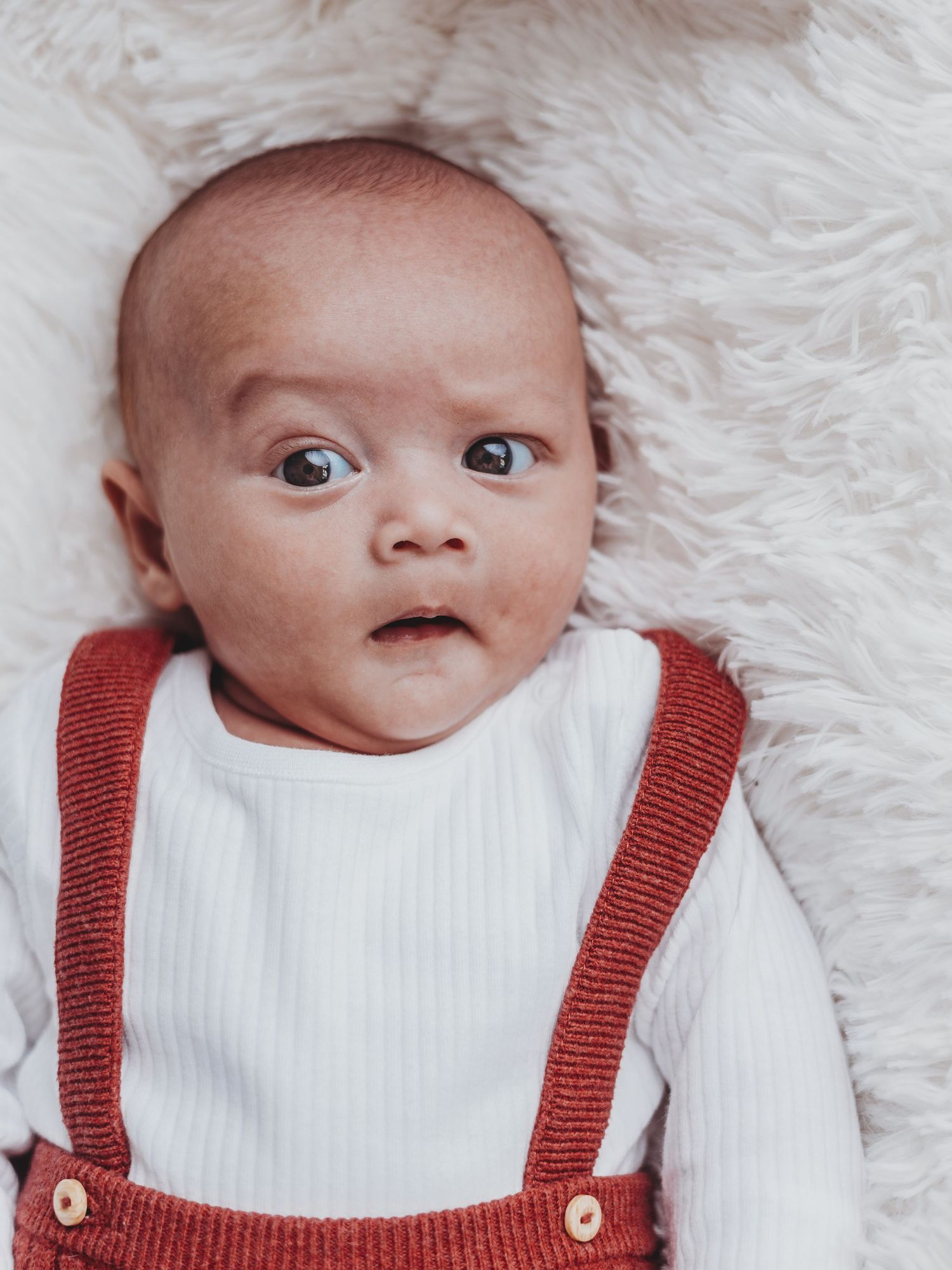 A baby in white and red overalls lying on a fluffy white blanket makes different adorable facial expressions.