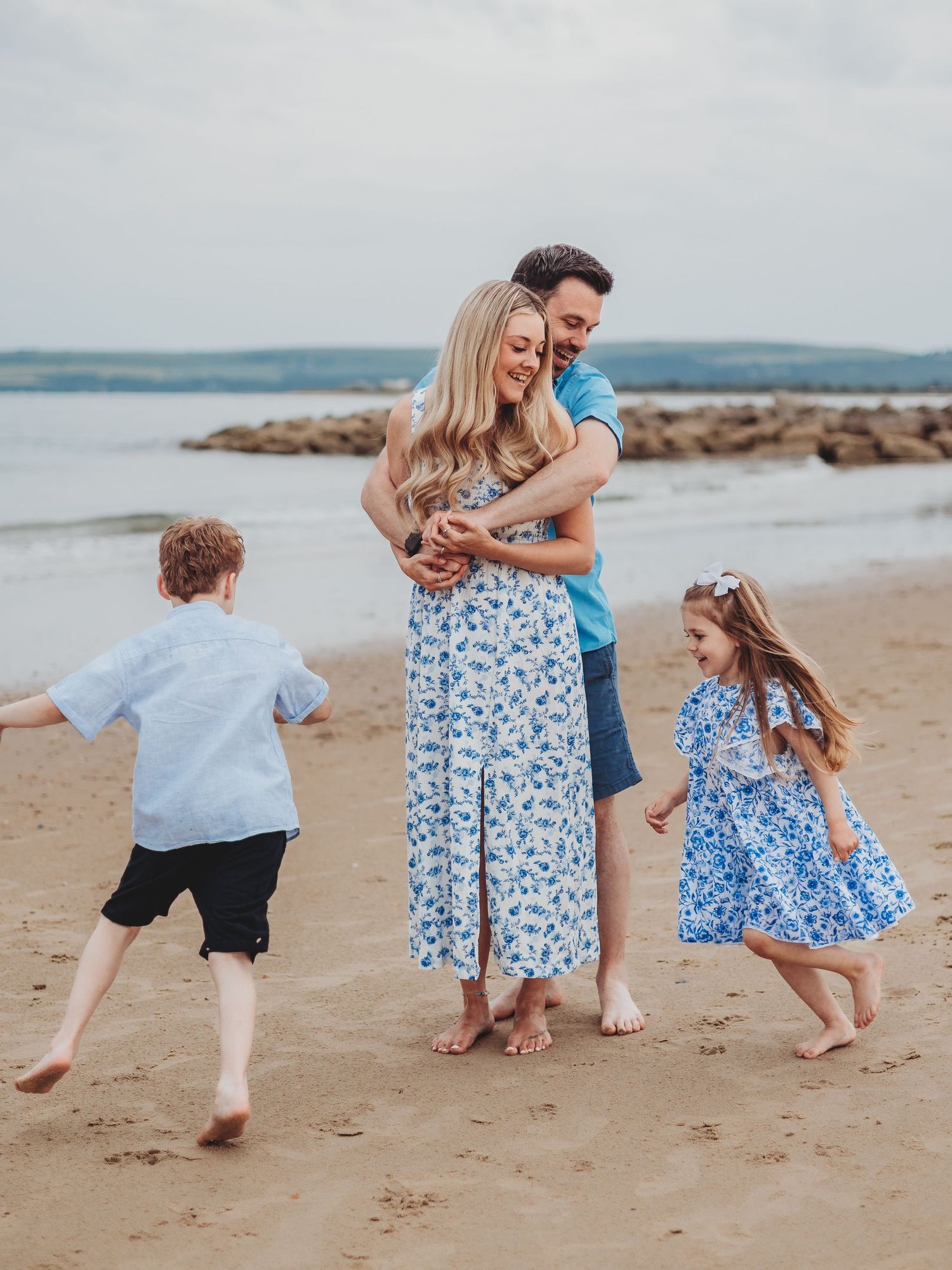 Family of four enjoying a beach day, parents embracing while two young children play on sandy shore.