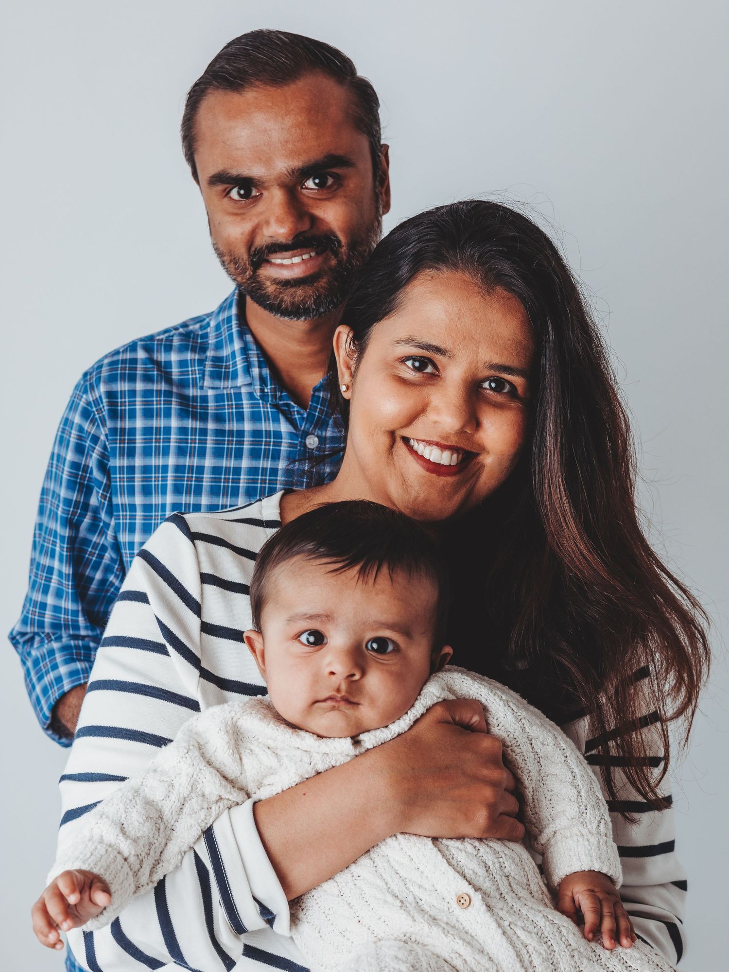 A smiling South Asian family with a man in a blue plaid shirt, a woman in stripes, and their baby in a white sweater.