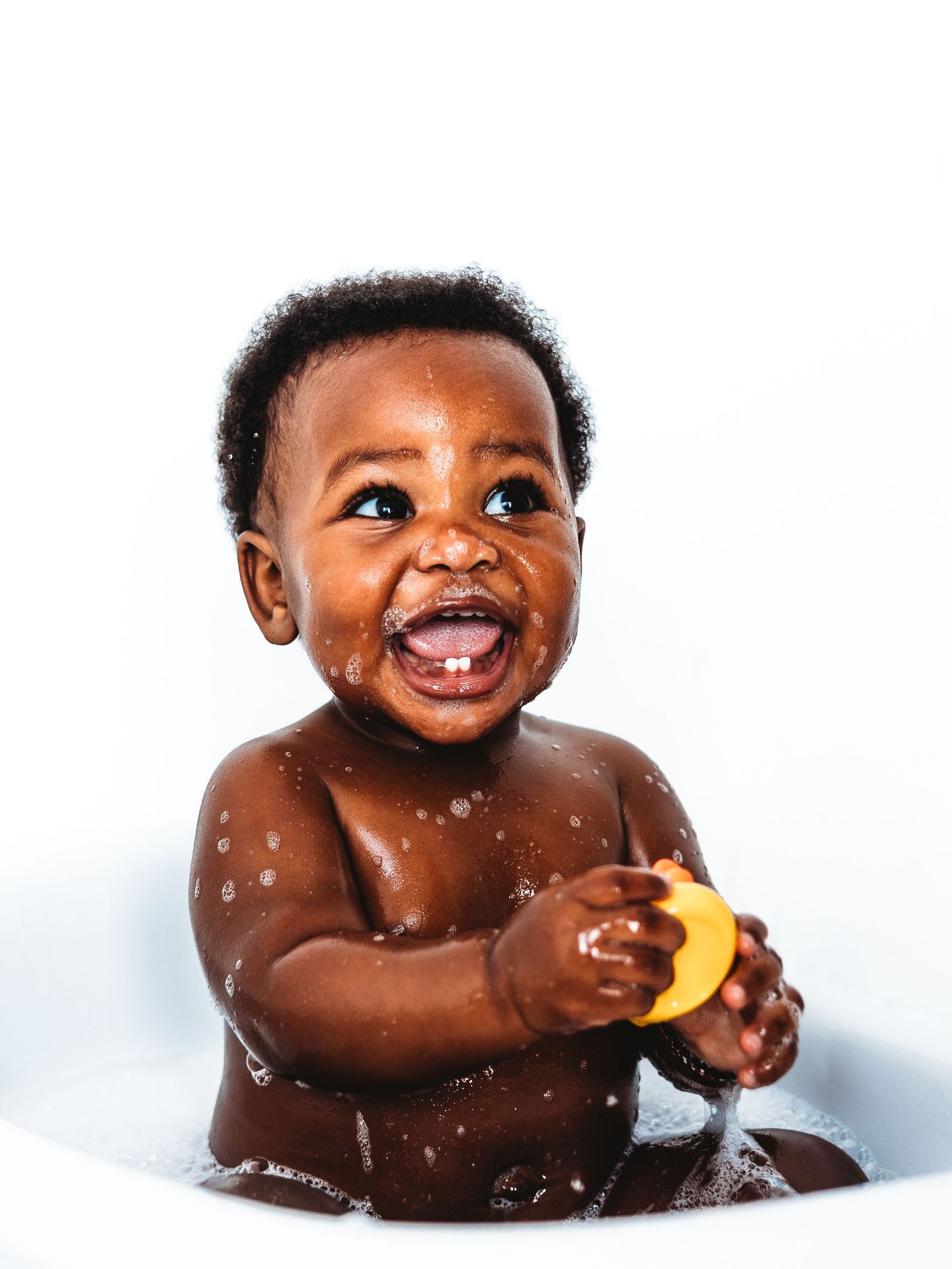 Smiling baby enjoying a bath, holding a yellow rubber duck, with water droplets on skin against white background.