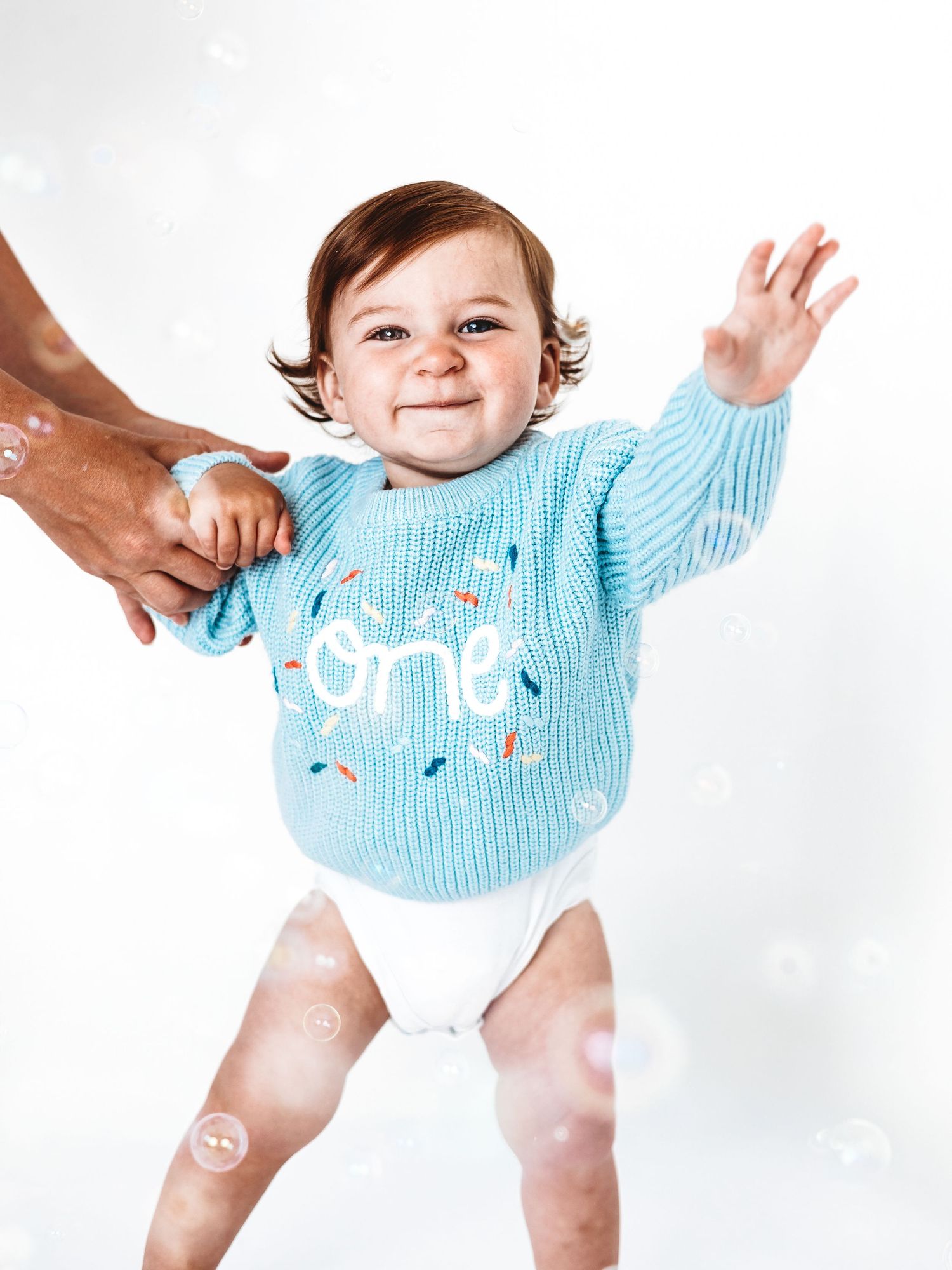 Smiling baby wearing a blue 'one' birthday sweater and diaper, standing with help, surrounded by bubbles.