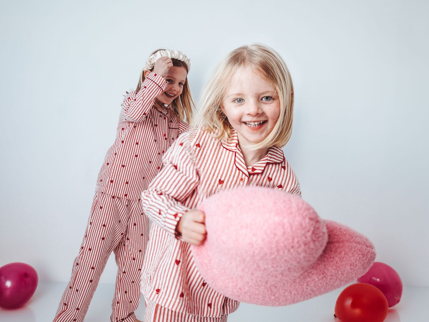 Two girls in pink striped and heart-print pajamas pose playfully with a pink heart pillow and balloons.