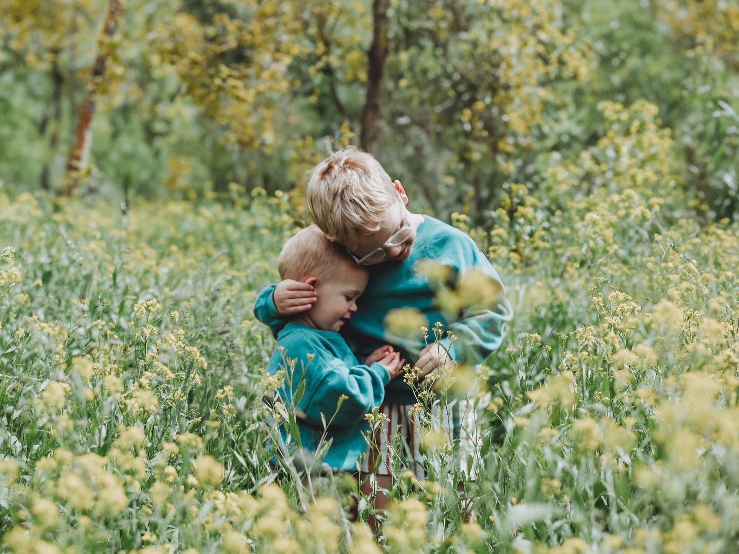 Two young children hugging in a vibrant field of yellow wildflowers surrounded by lush green trees.