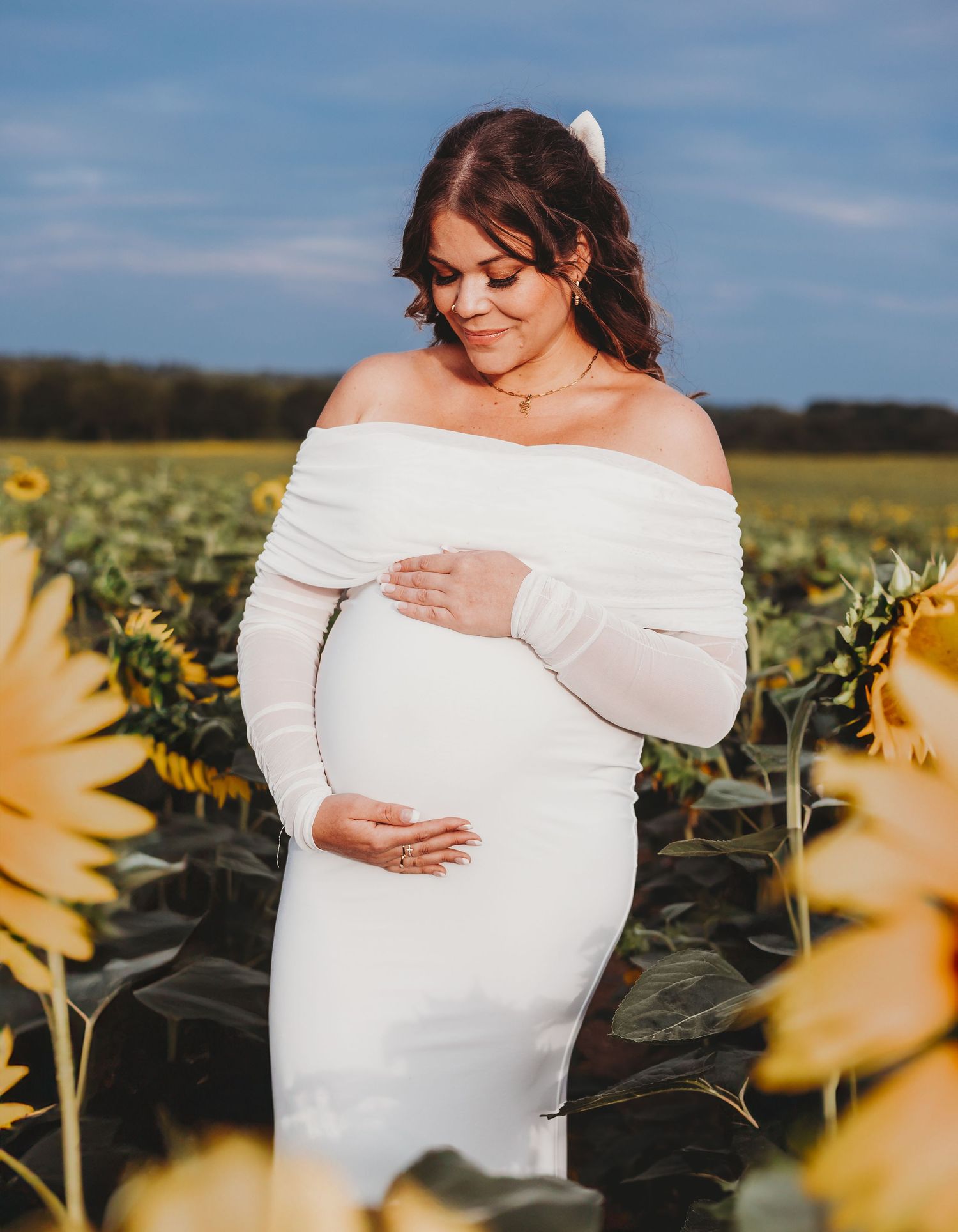 A maternity photo shows an off-shoulder white dress against a vibrant sunflower field backdrop on a sunny day.