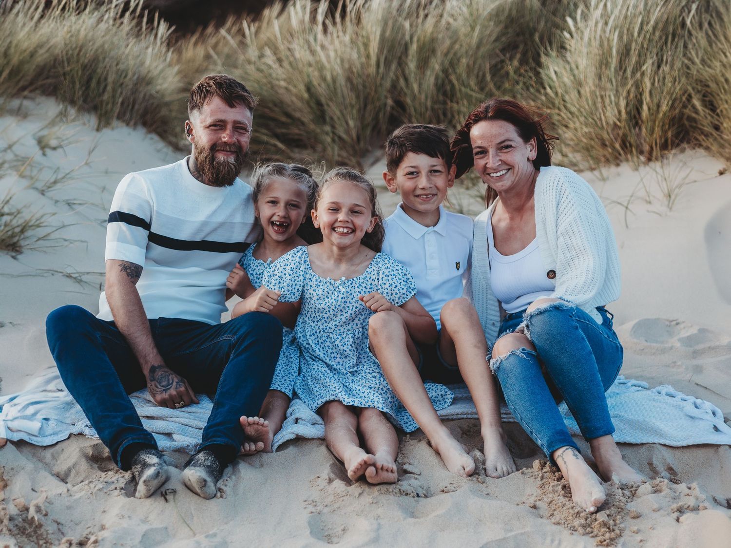 Happy family of five smiling together on a sandy beach, sitting on a blanket near grass-covered dunes.