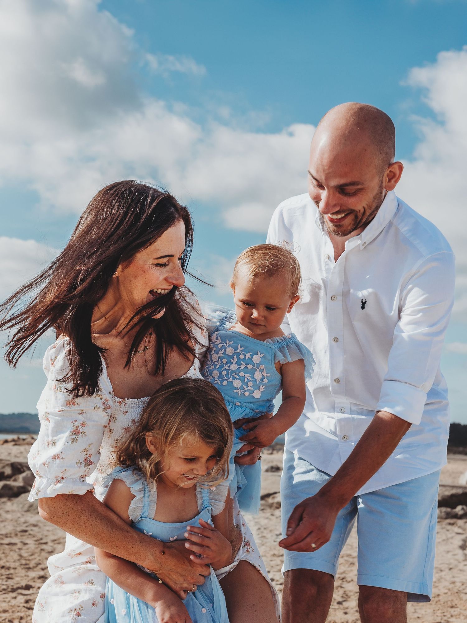 Happy family of four enjoying a sunny beach day, parents smiling at two young children dressed in blue and white.