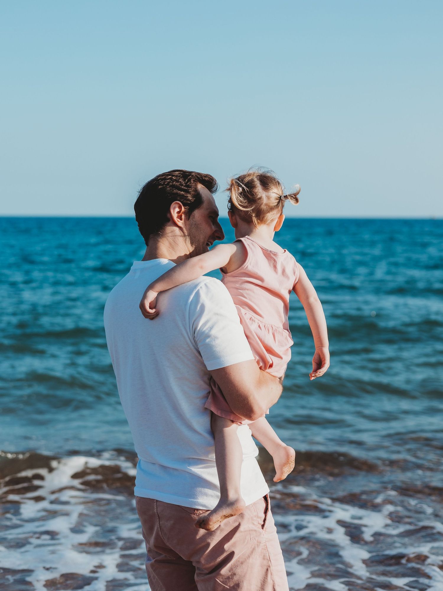 Man holding young child at beach with ocean waves and clear blue sky in background.