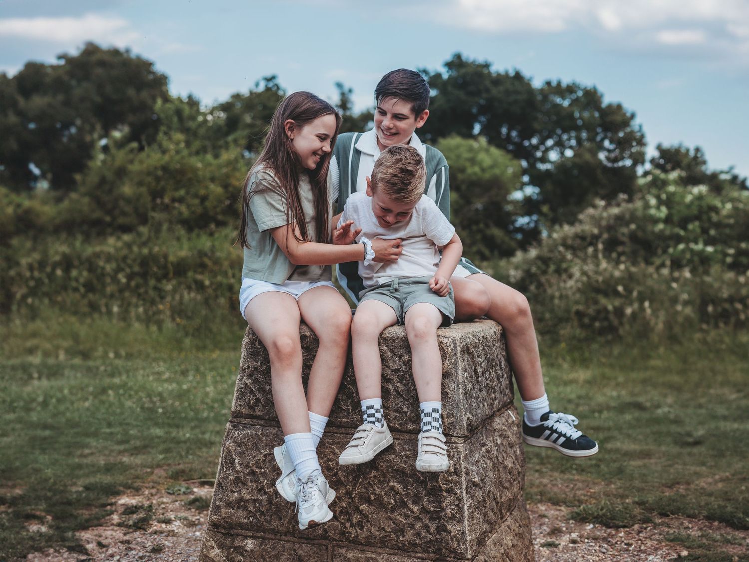 Three children sitting on a stone block outdoors, smiling and looking at a phone in a green park setting.