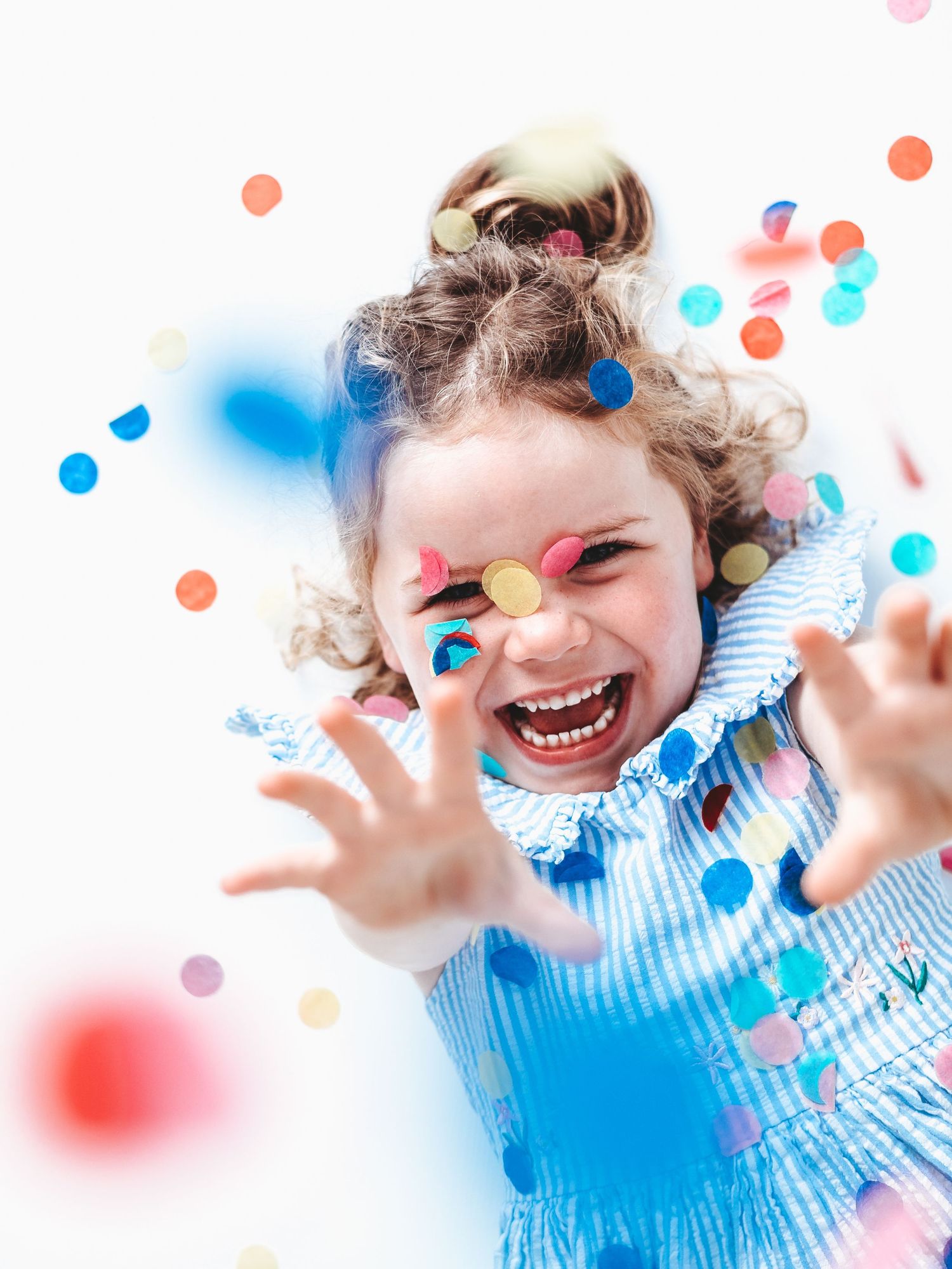 A joyful young girl in a blue dress laughs and reaches toward colorful confetti falling around her.