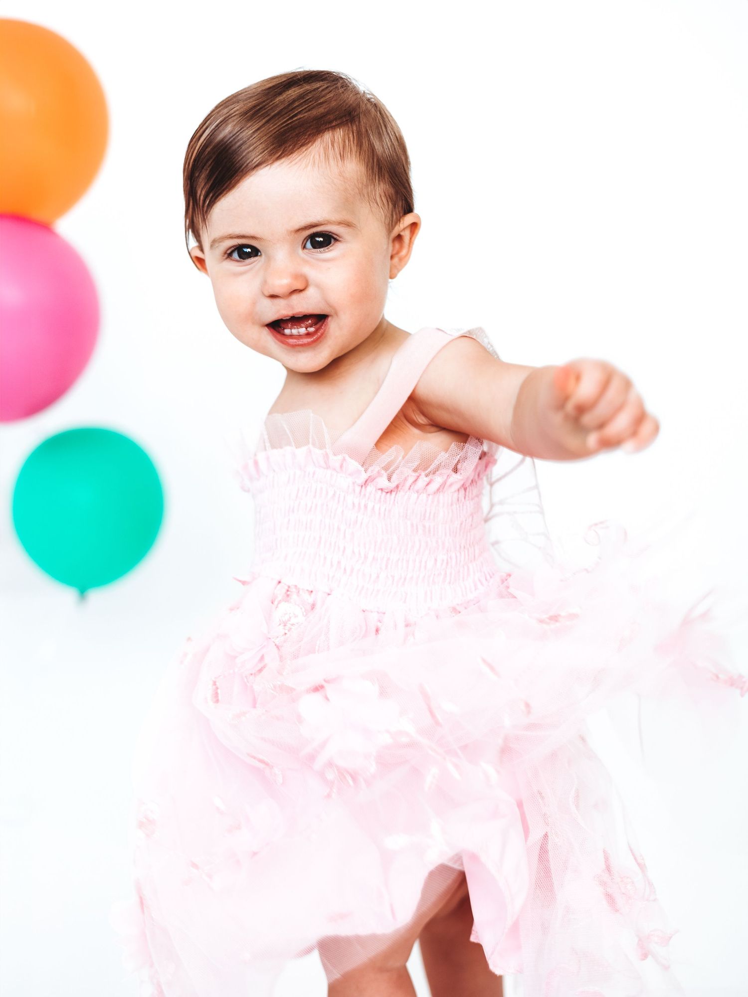A smiling baby girl in a pink tutu dress reaches out joyfully, surrounded by colorful balloons on a white background.