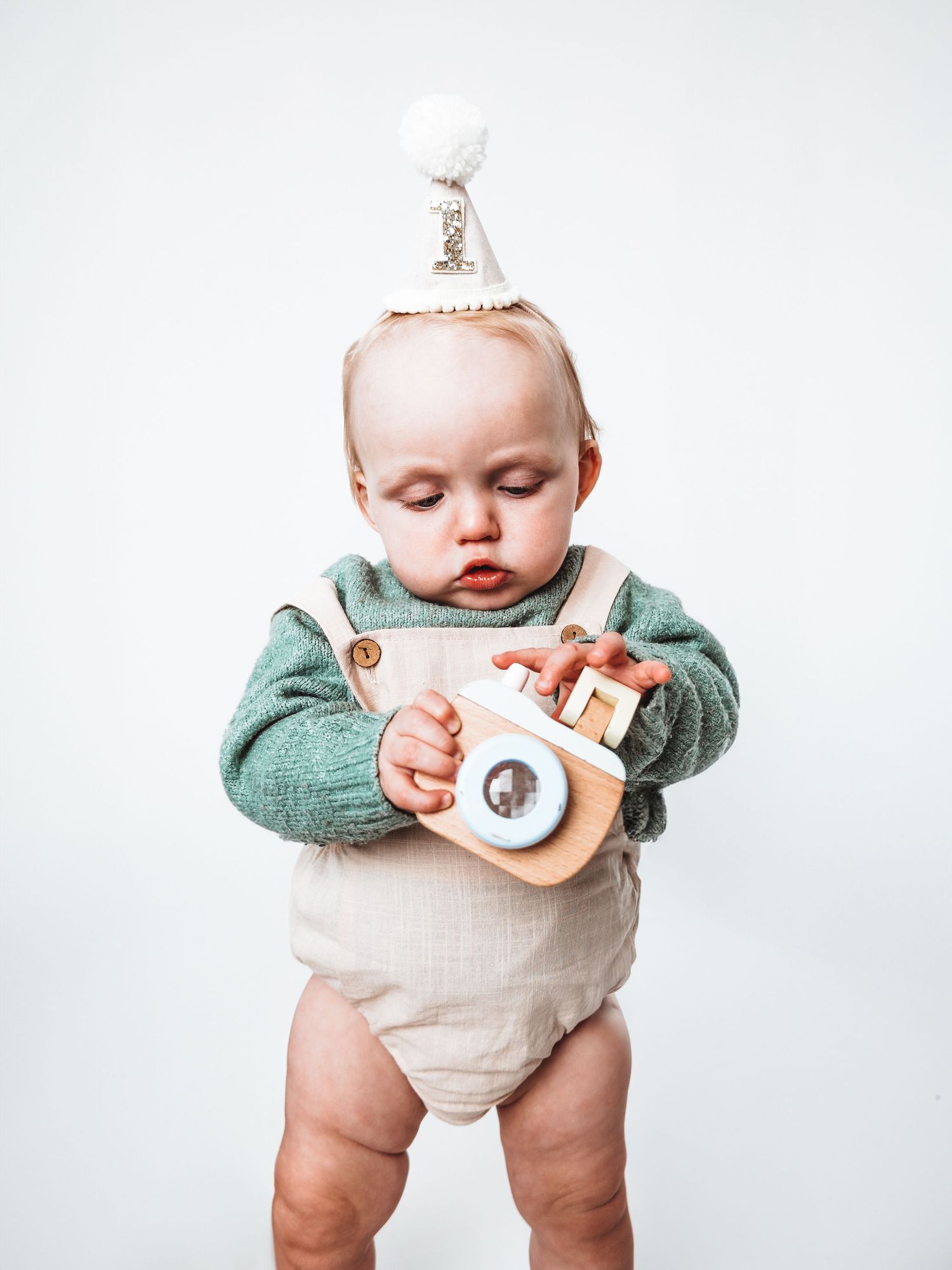 A cute baby wearing a white party hat and green sweater romper holds a wooden toy camera against a white background.