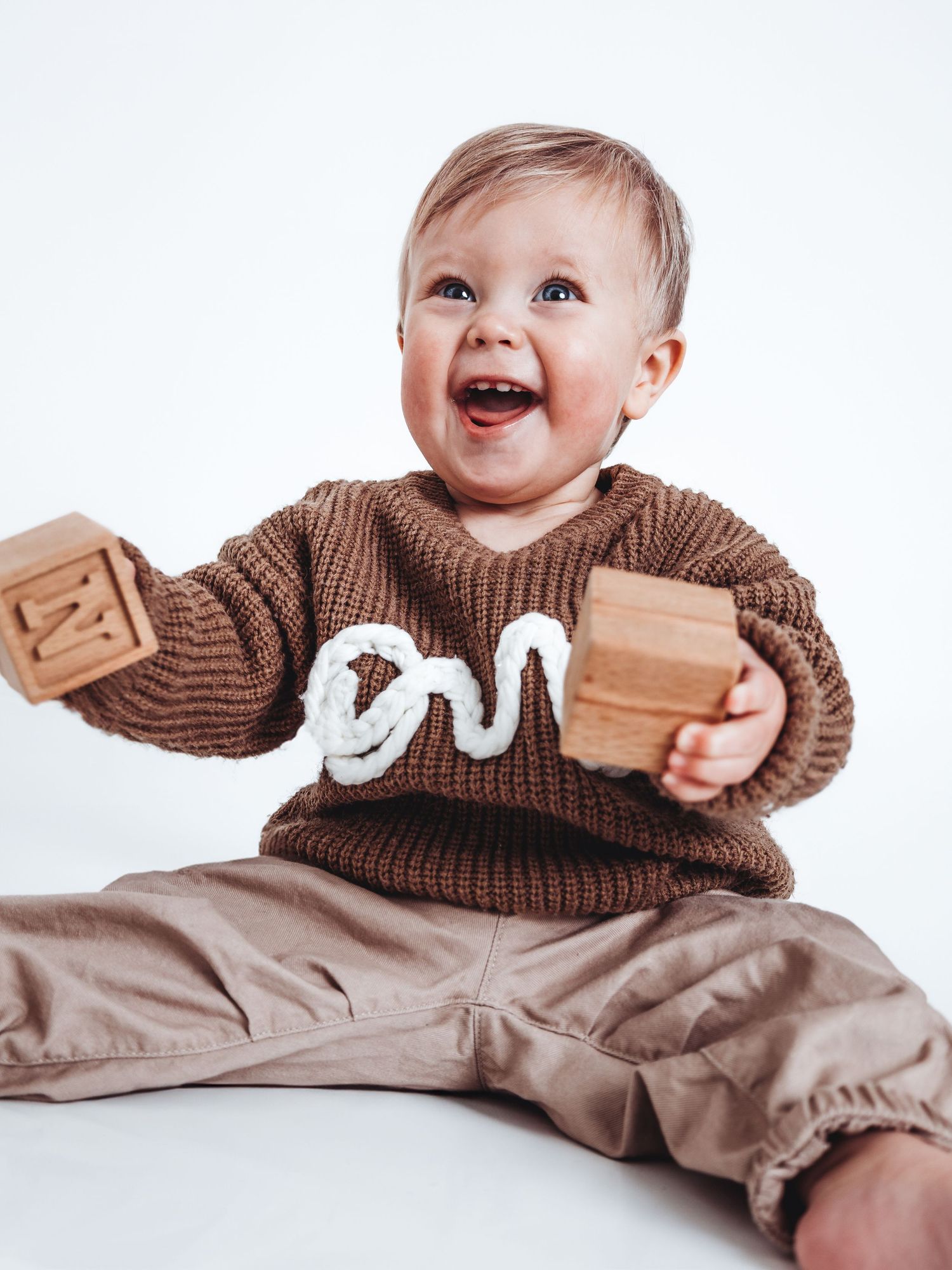 Happy baby in brown knit sweater holding wooden alphabet blocks, sitting against white background.