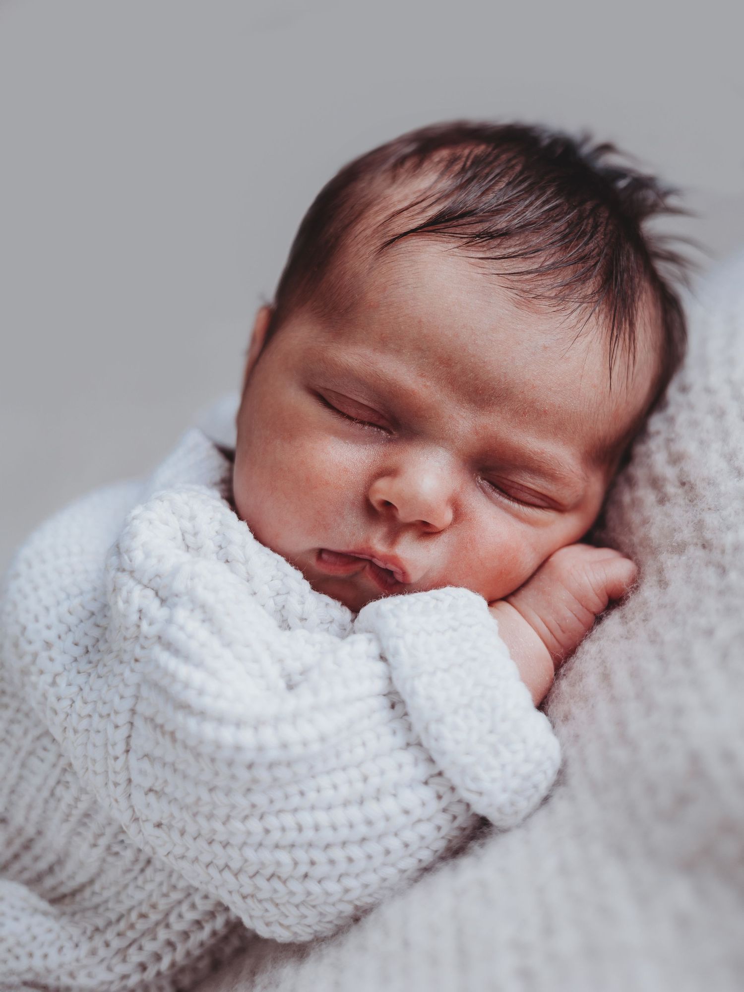 A sleeping newborn baby wrapped in a cozy white knit sweater rests peacefully against a soft background.