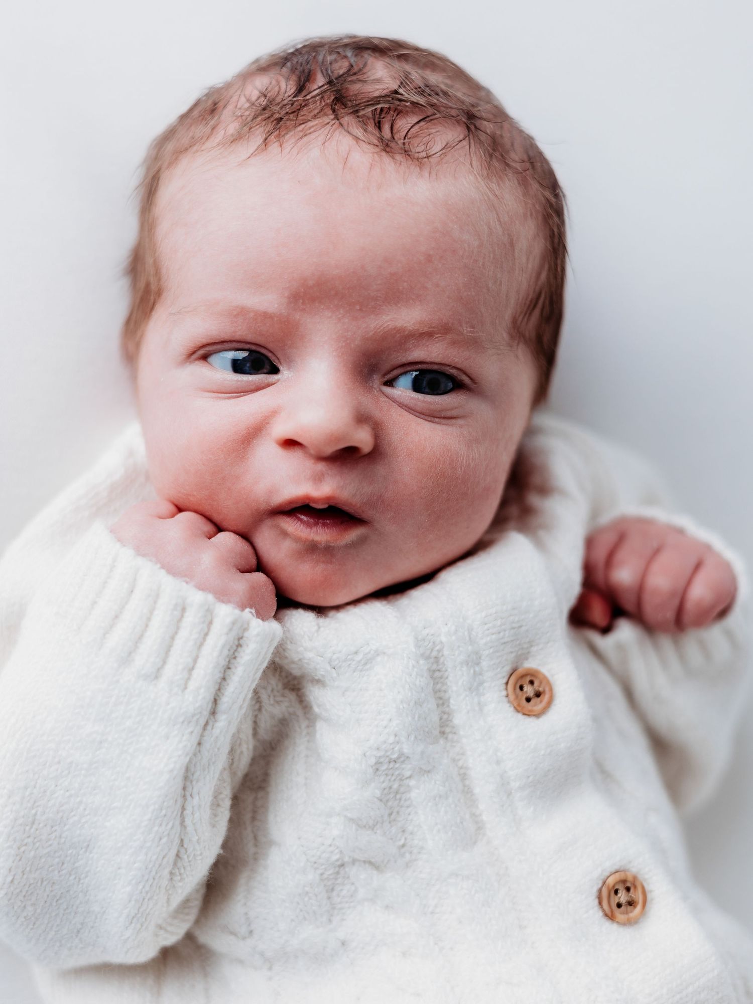 A small baby wearing a cozy white knit sweater with wooden buttons smiles while lying down.