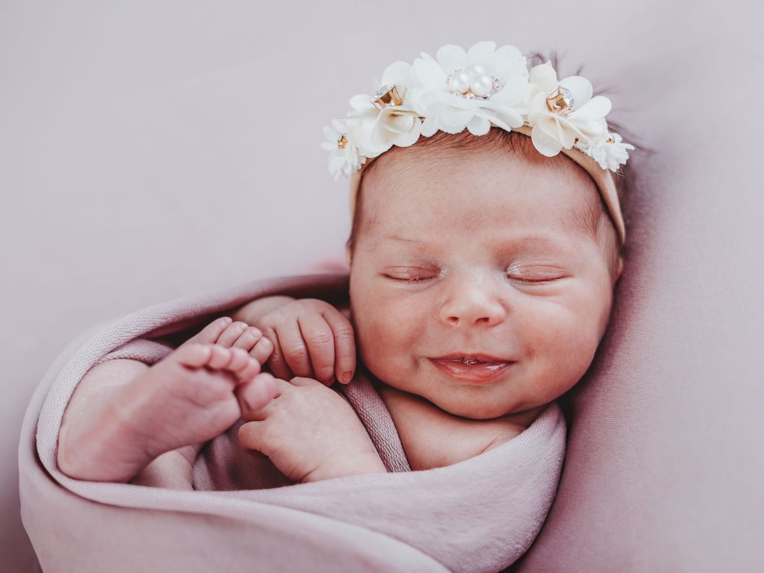 Sleeping newborn baby wearing white floral headband wrapped in soft pink blanket.