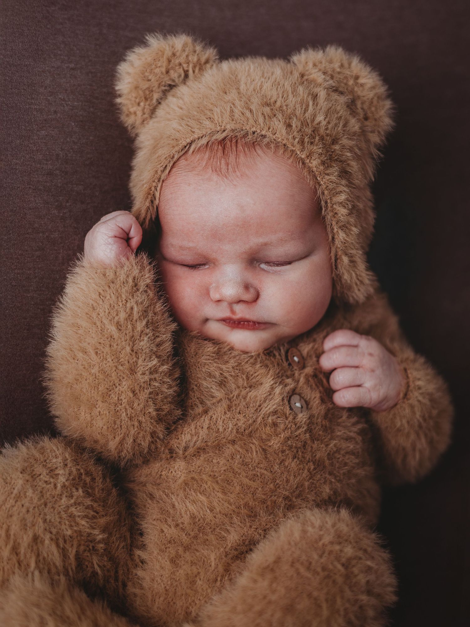 A sleeping baby wearing a cozy brown teddy bear costume with fuzzy ears lies peacefully against a dark background.