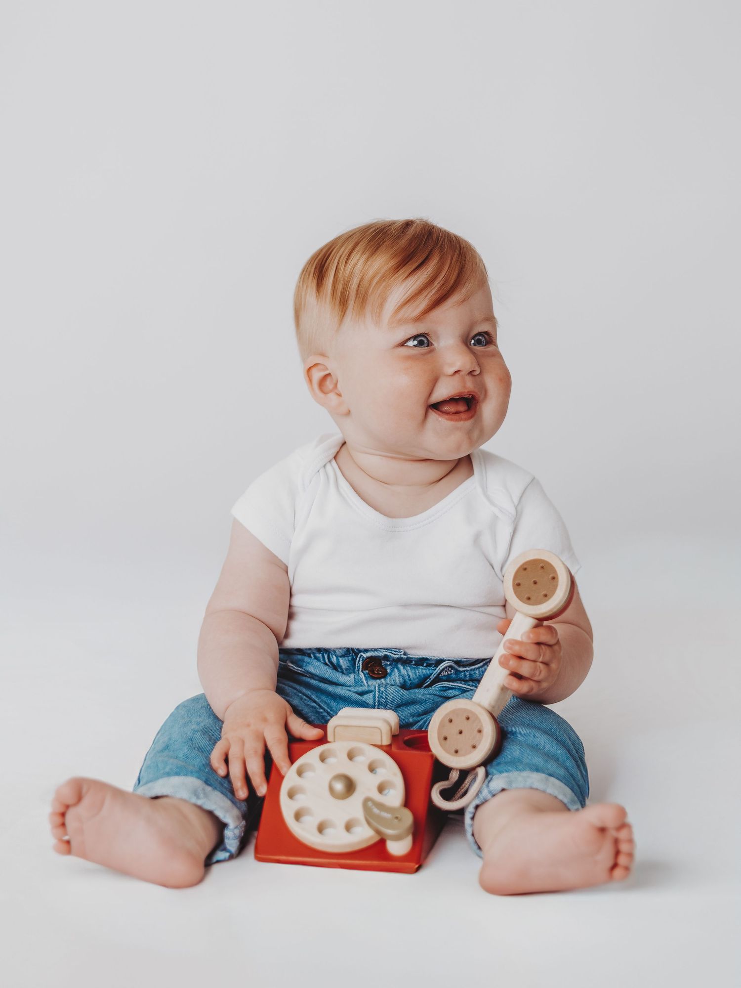 Baby in white shirt and jeans sits on white background holding wooden toys and smiling with a red toy phone.