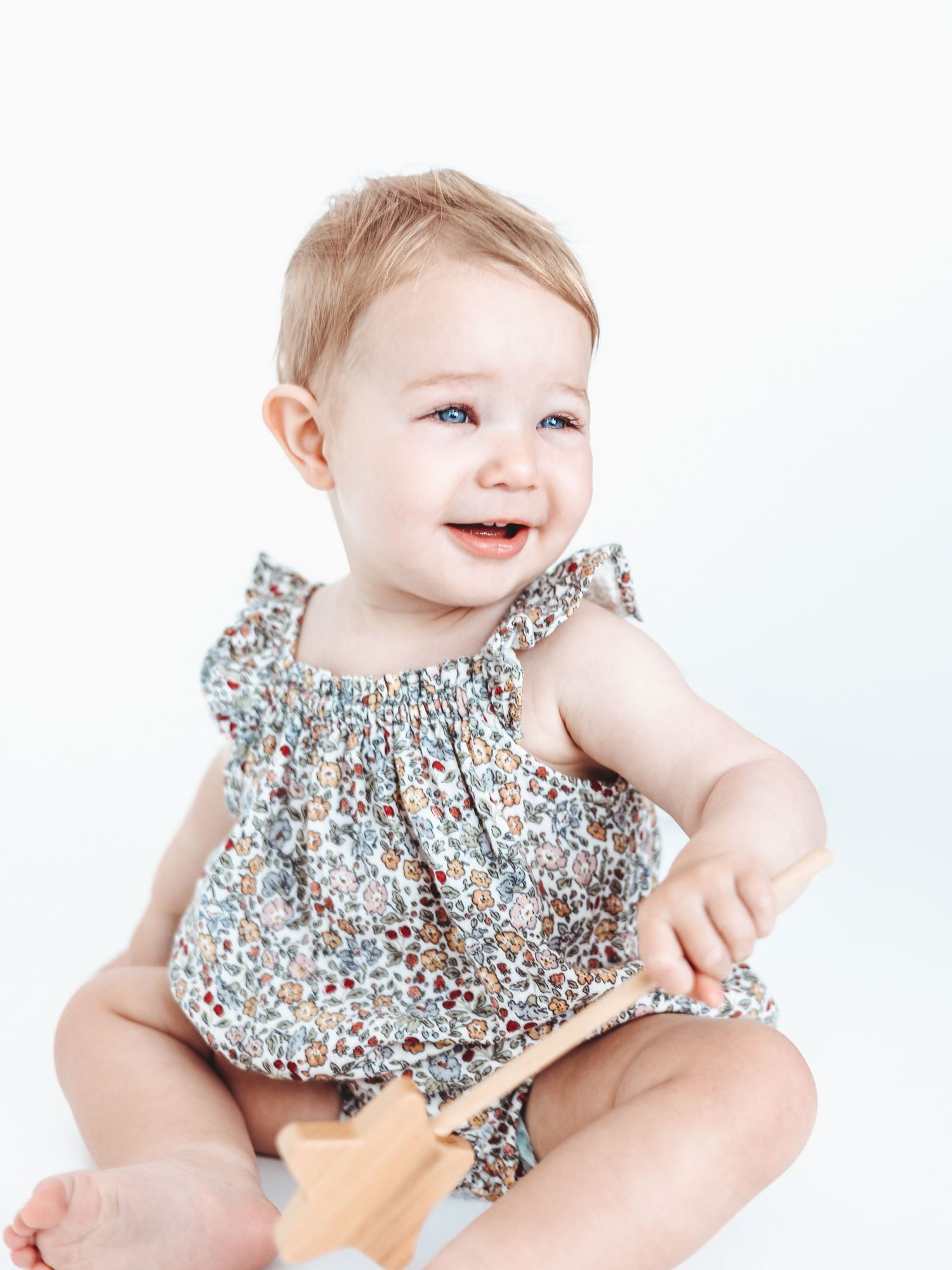 Baby wearing floral print ruffled romper smiles while sitting against white background.