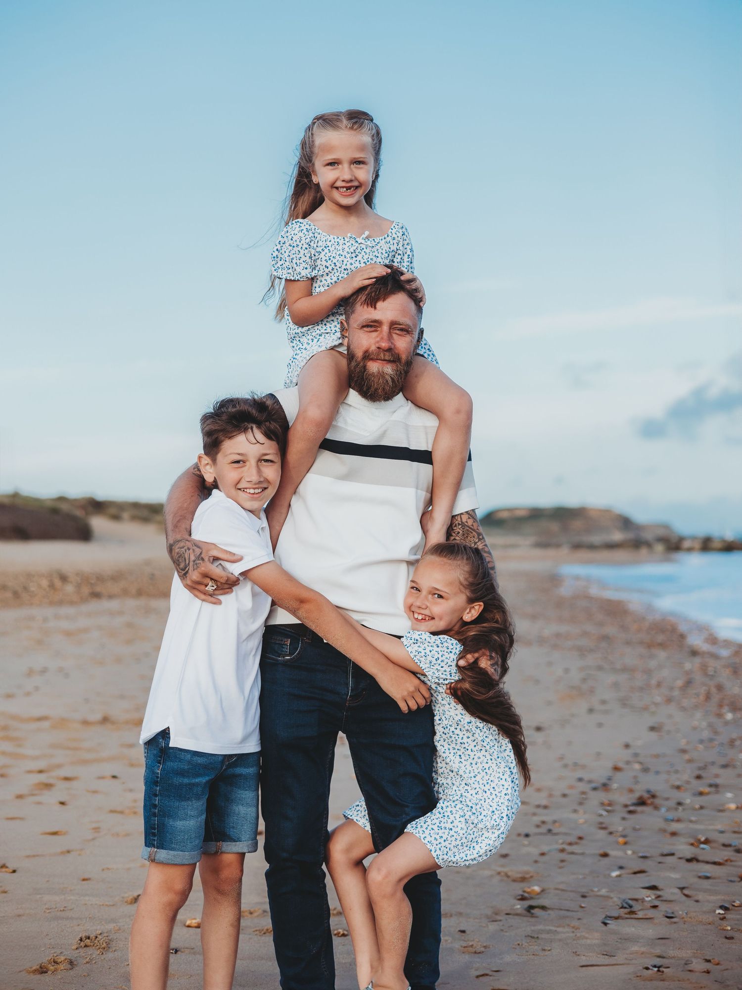 Bearded father poses on beach with three children, one on shoulders and two hugging him, all smiling.