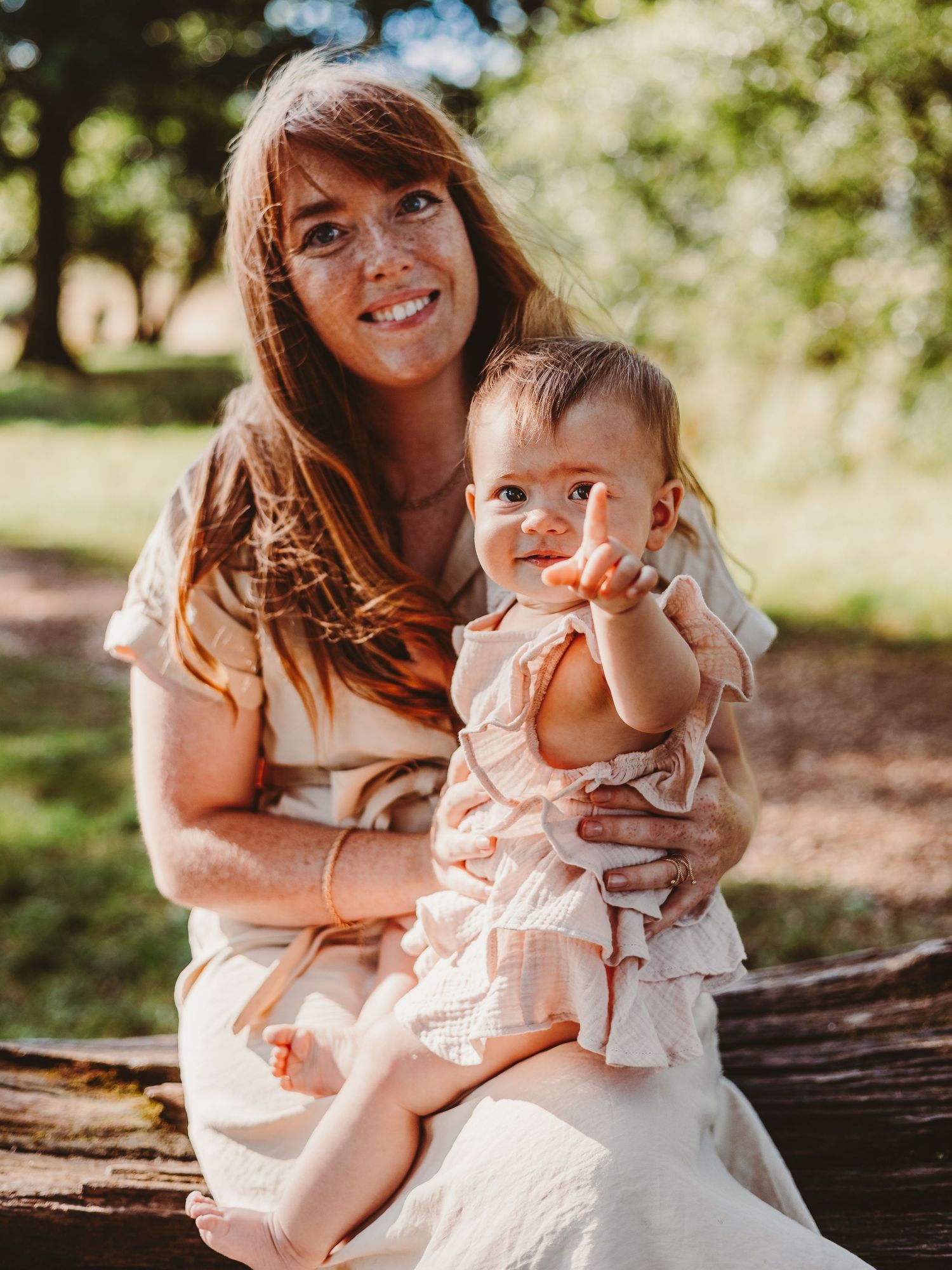 A smiling woman with long red hair holds a happy baby girl in a pink dress outdoors in a lush green park.