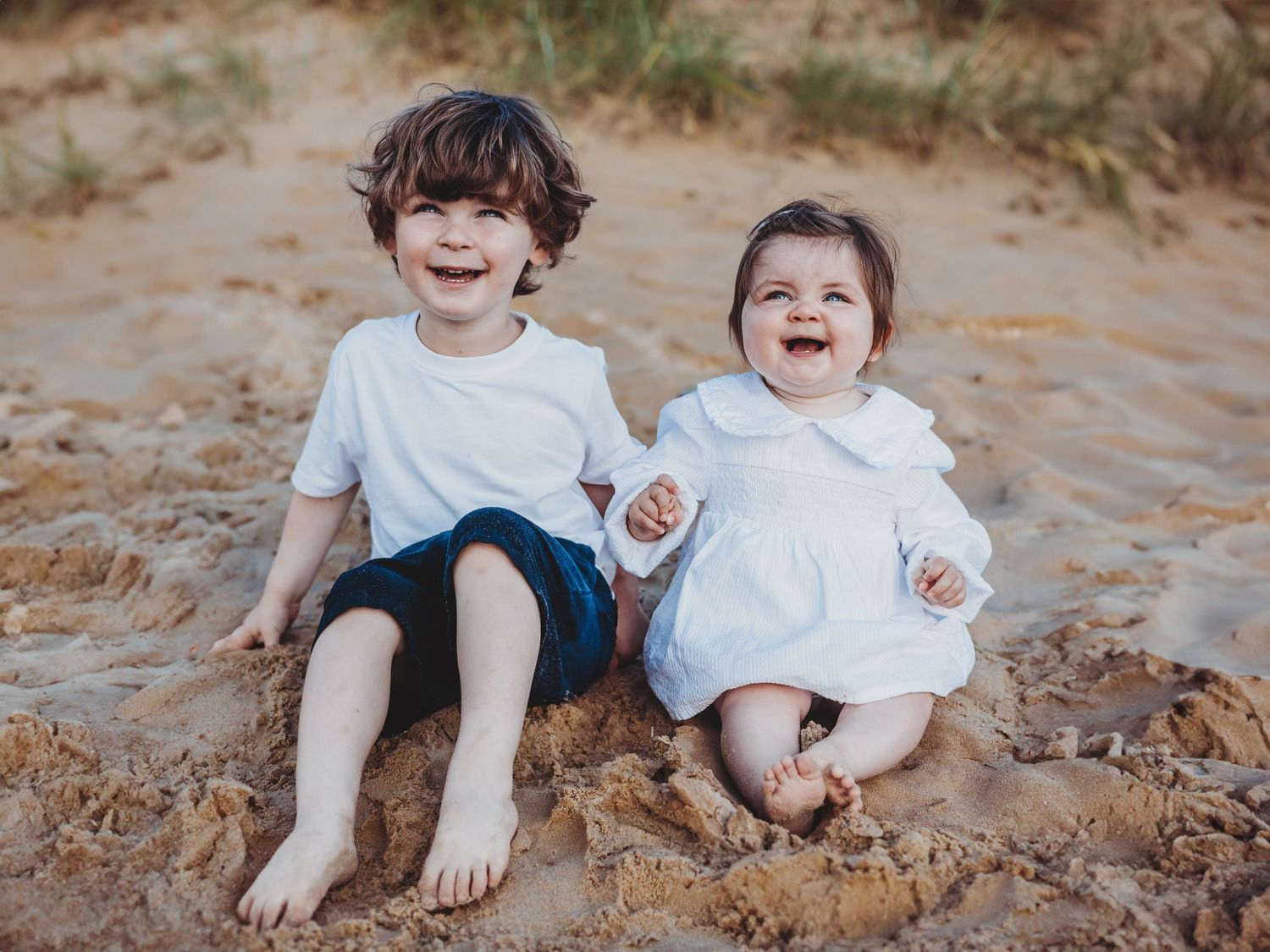 Two happy children sitting on sandy beach, smiling boy in white tee and navy shorts beside laughing baby girl in white dress.