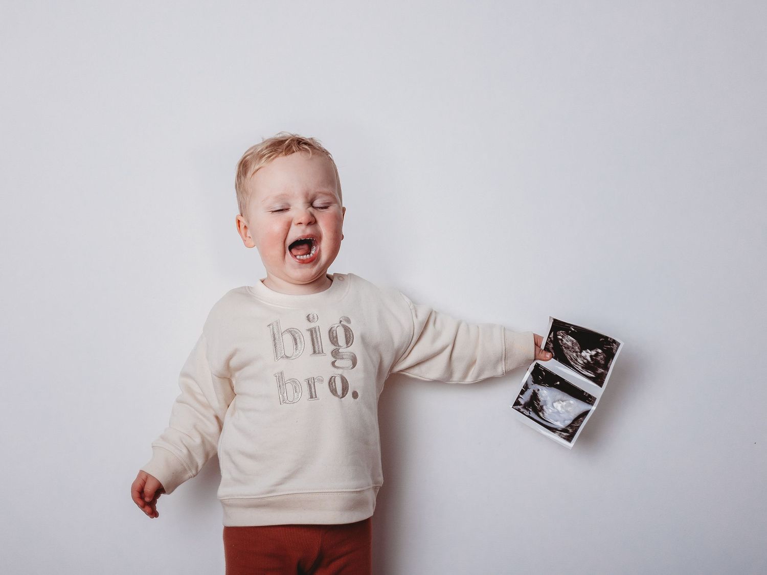 Excited toddler in a big bro sweatshirt holding ultrasound photos announcing a new sibling.