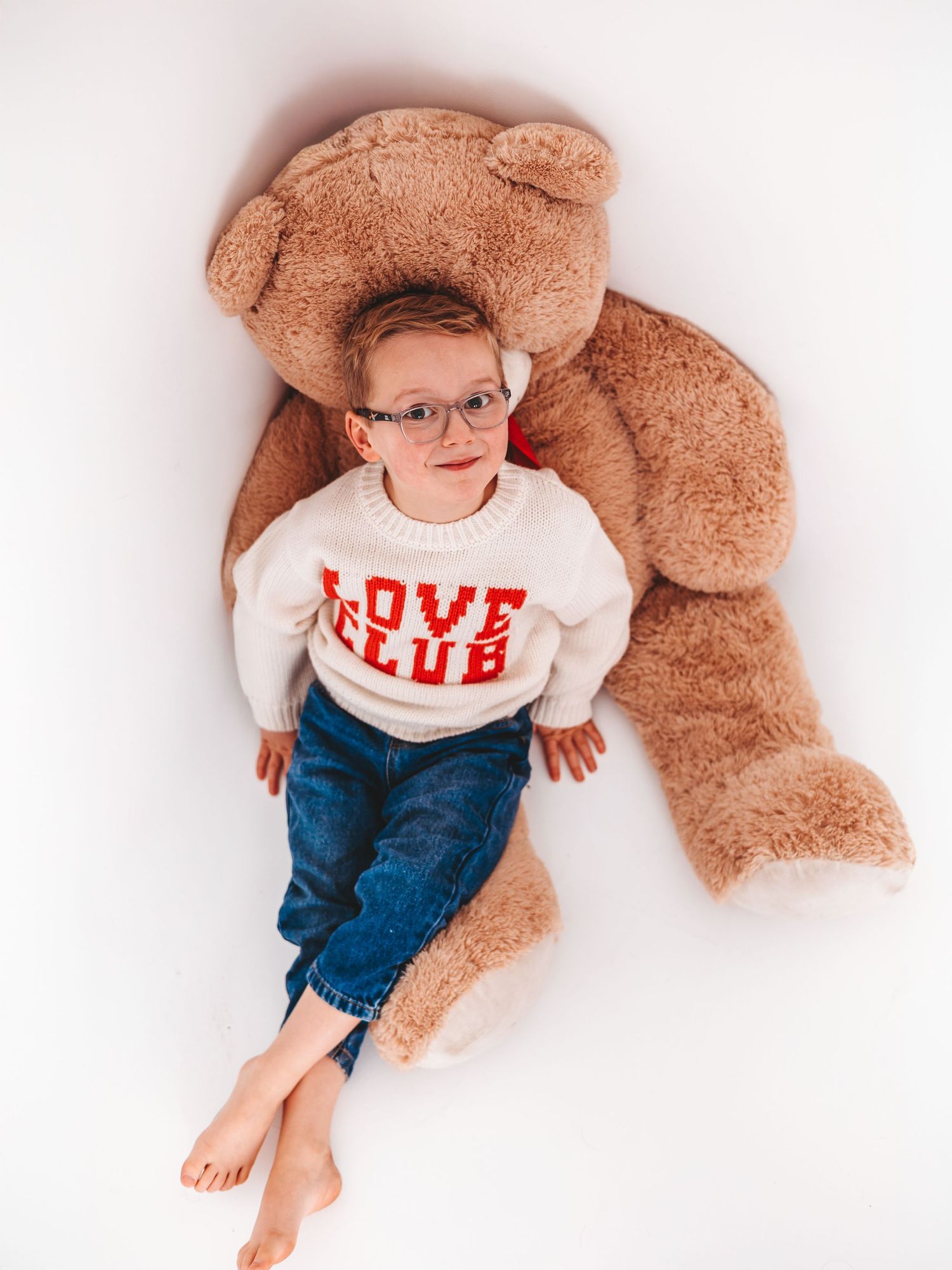 Young child in a white Love Club sweater leaning against a giant teddy bear on a white background.