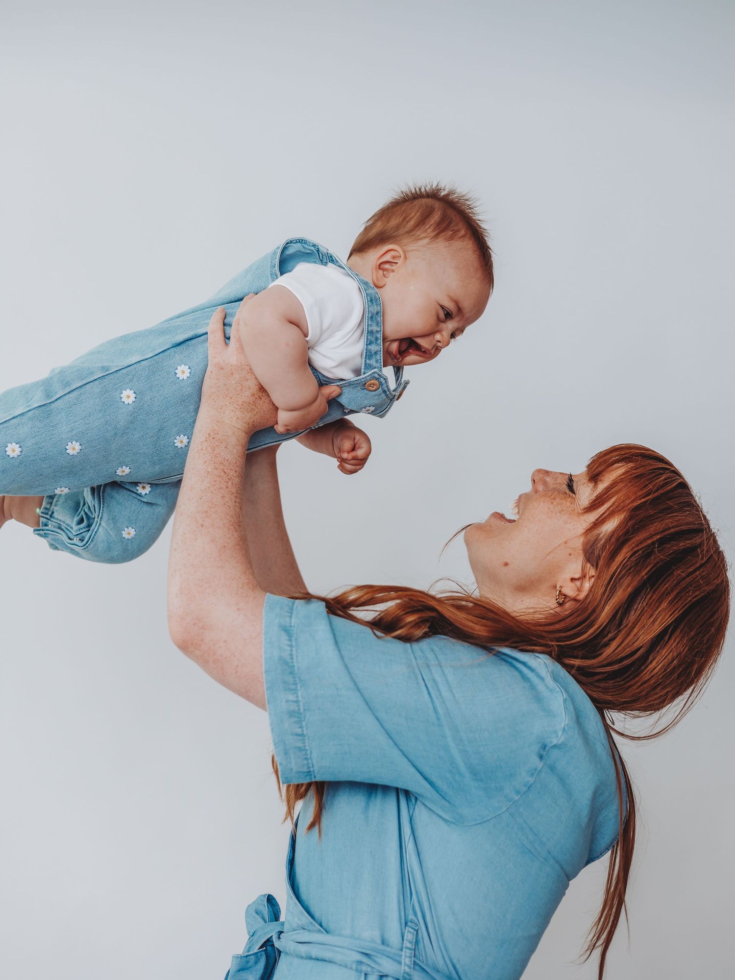 A joyful redhead mother in a blue dress lifts her smiling baby wearing a denim outfit against a light background.