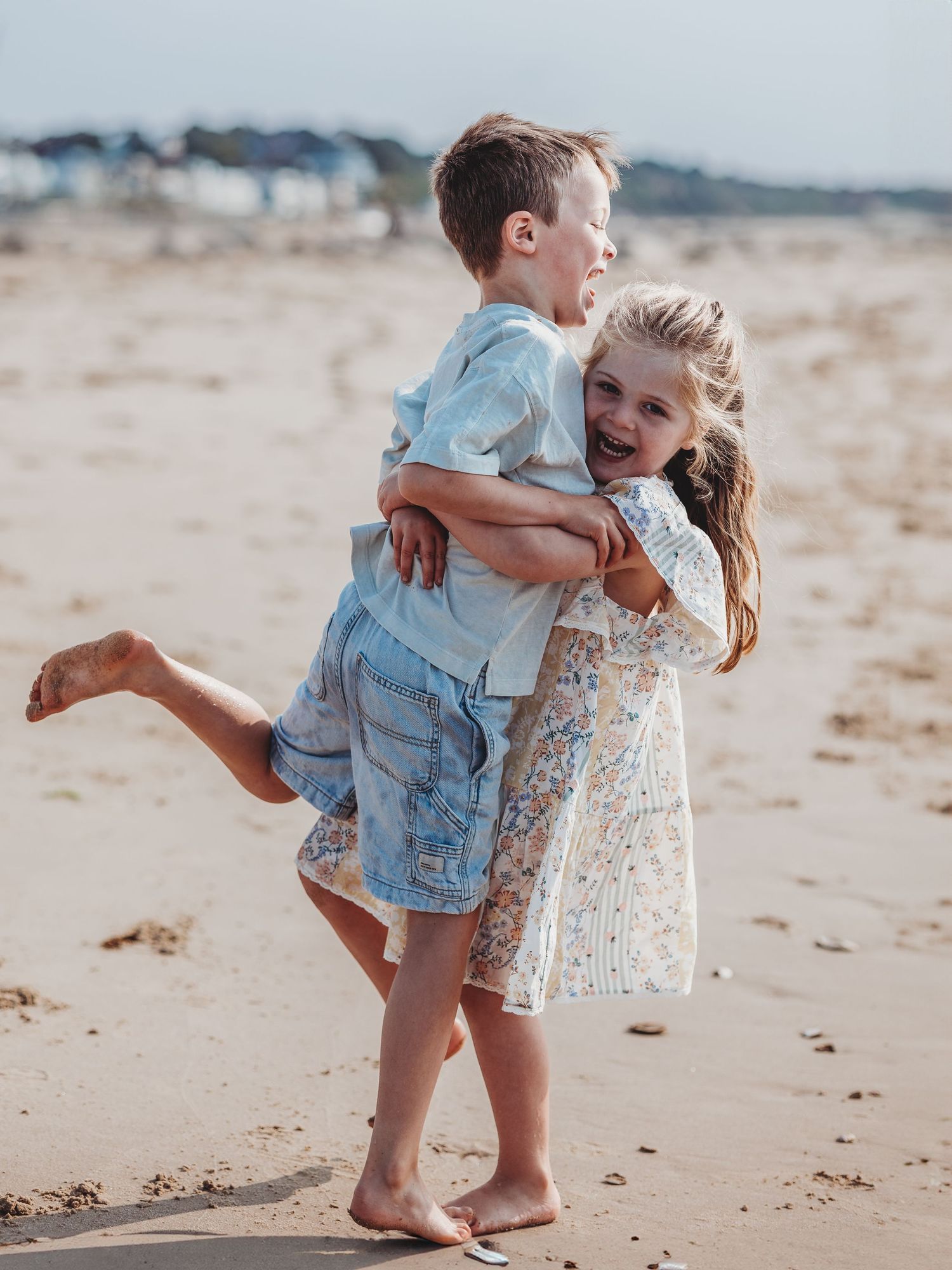 Two happy children playing on a sandy beach, with a girl hugging a boy from behind, both laughing and barefoot.