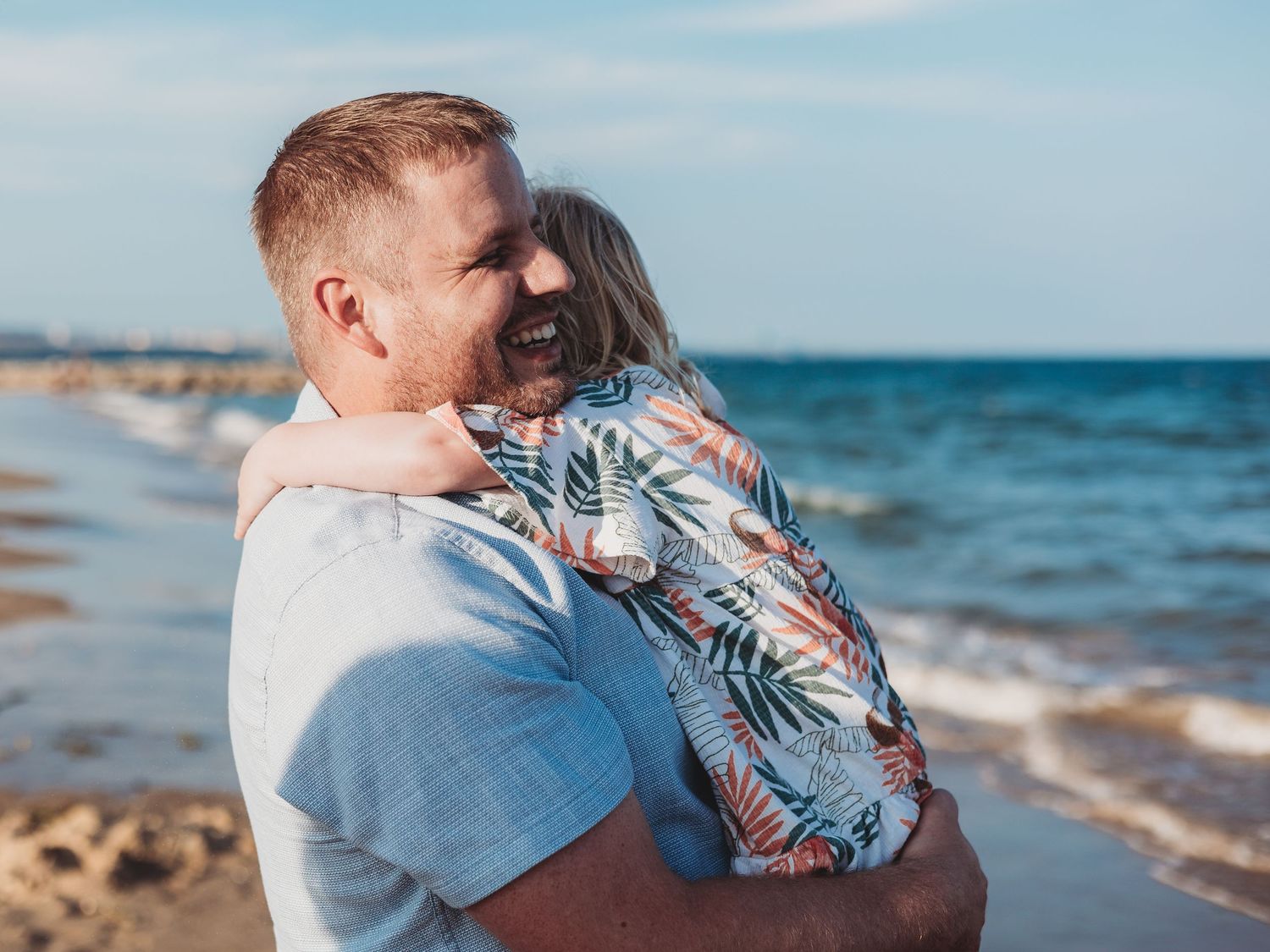 A father joyfully holds his laughing child on a sunny beach with ocean waves in the background.