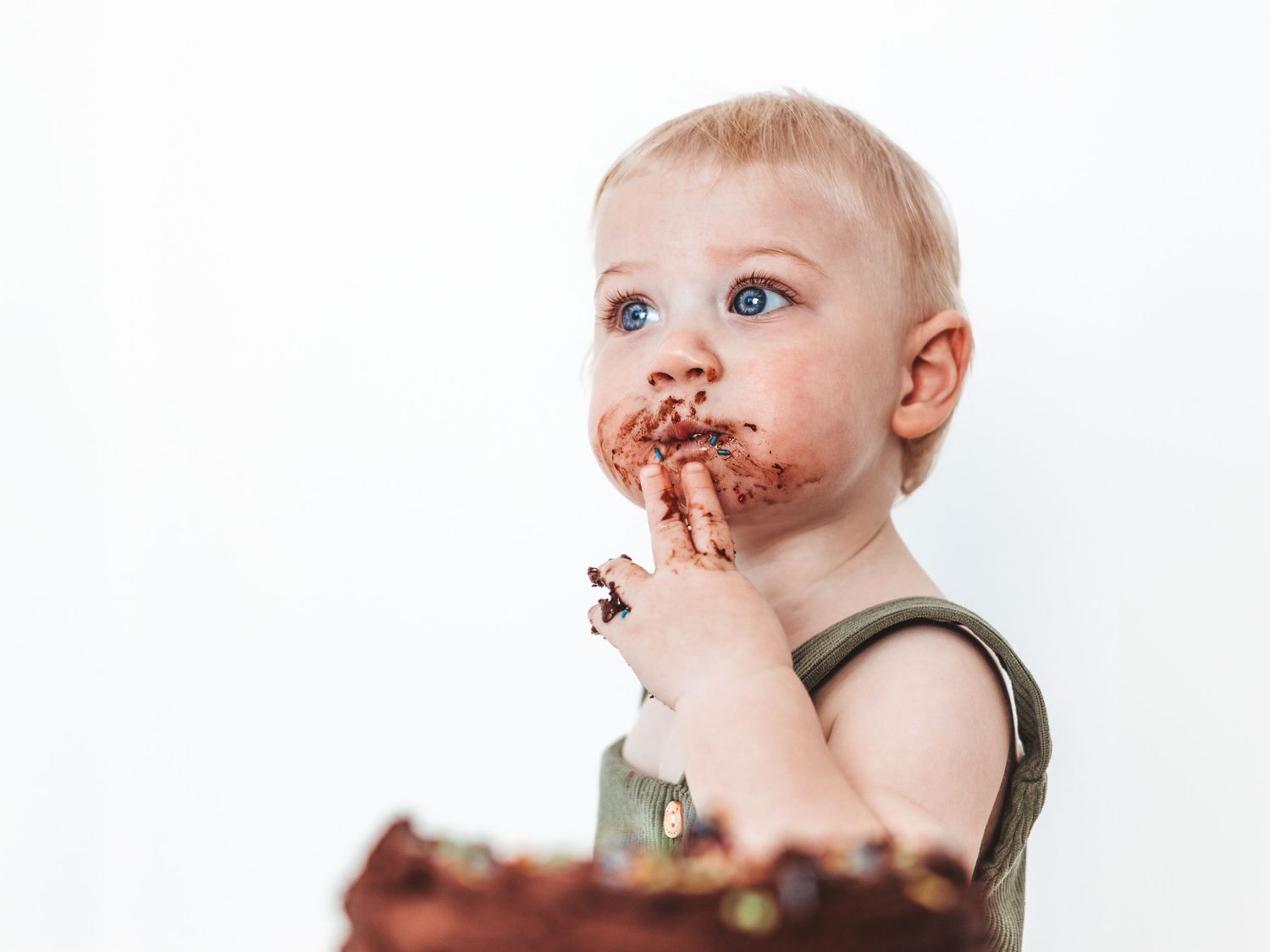 Toddler with blue eyes eating chocolate cake, face and hands covered in chocolate frosting, white background.