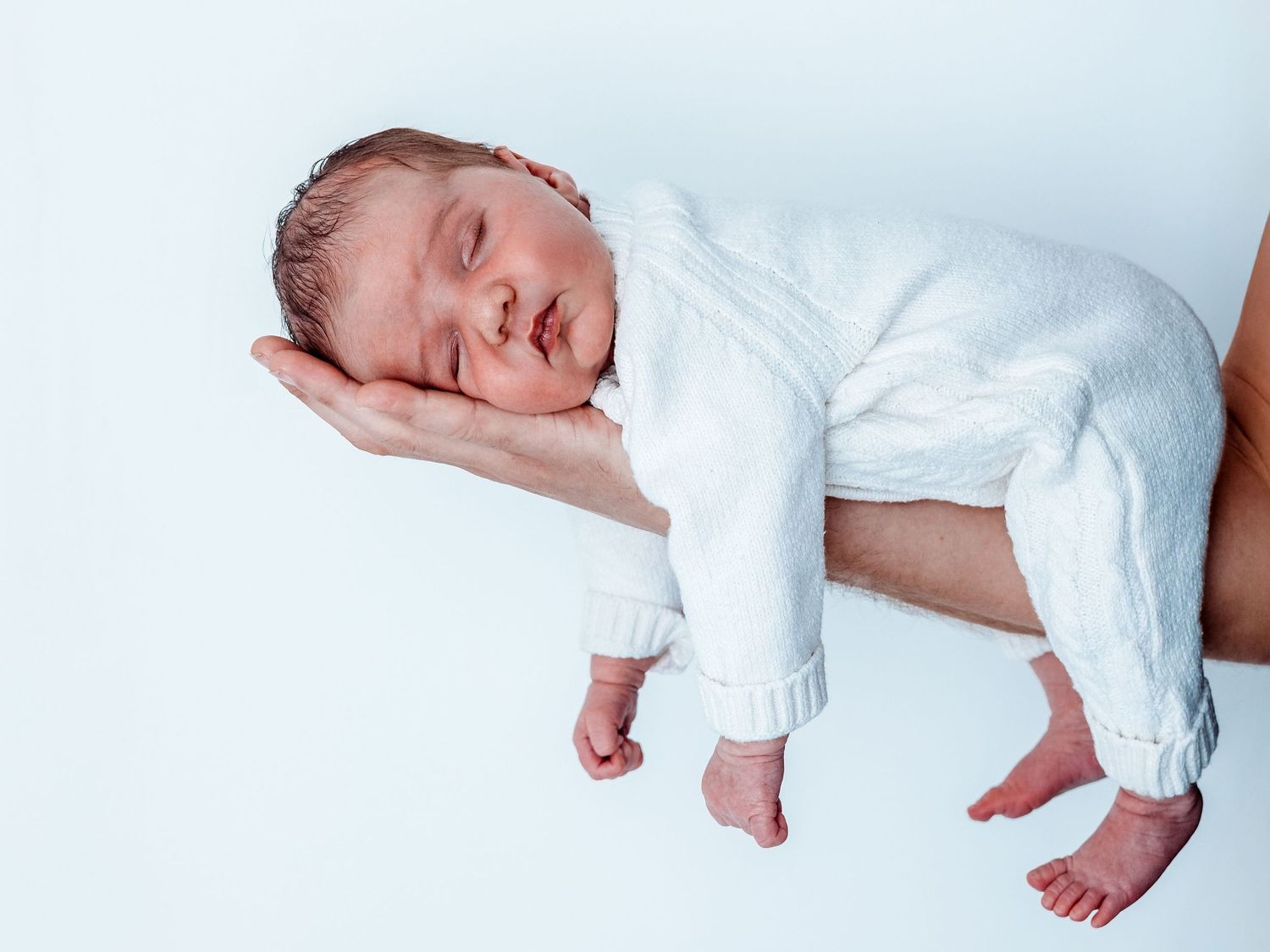 A sleeping newborn baby wearing white clothing lies peacefully on white bedding.