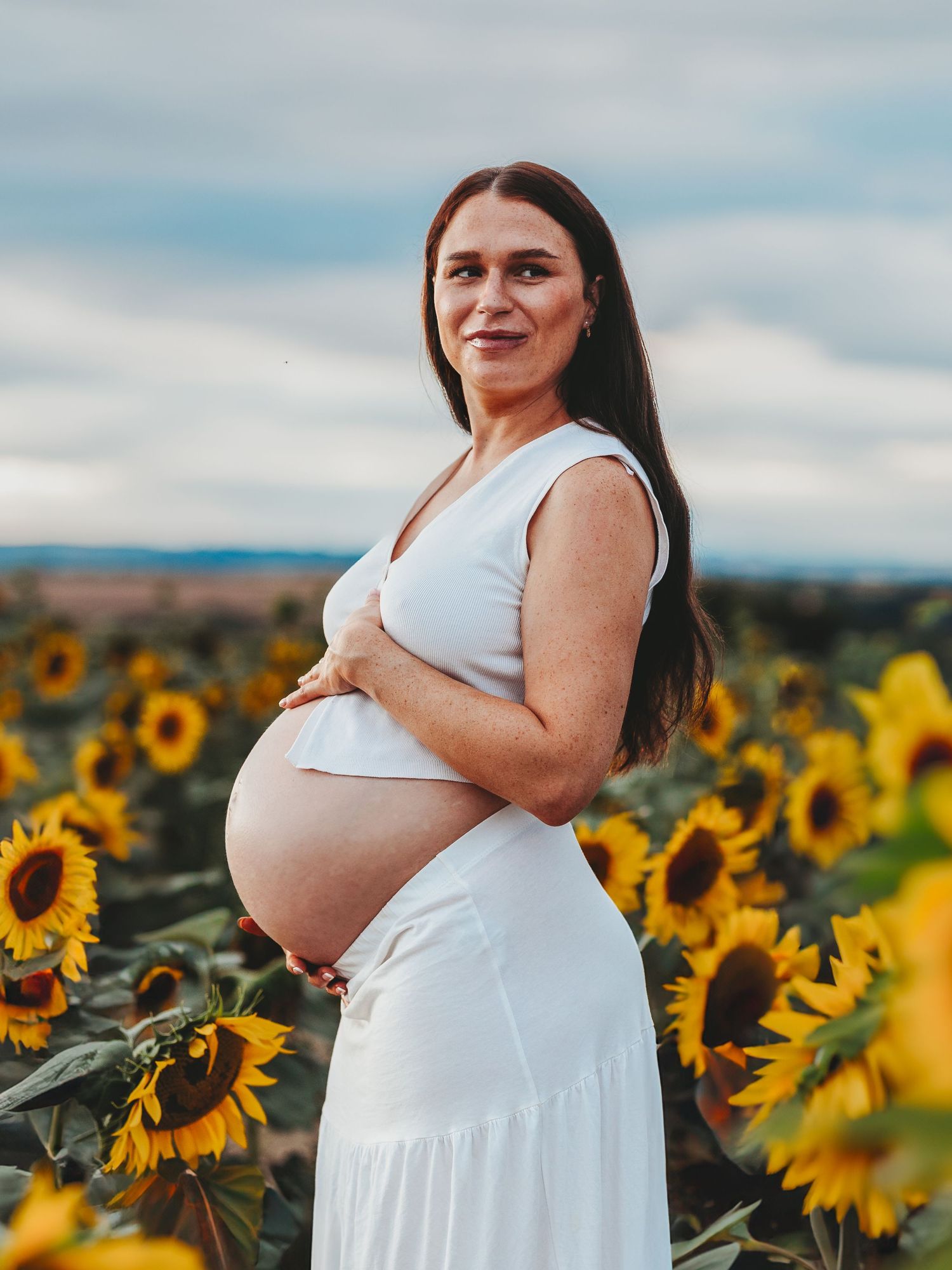 A person in a white dress poses in a vibrant sunflower field against a cloudy sky during a maternity photoshoot.