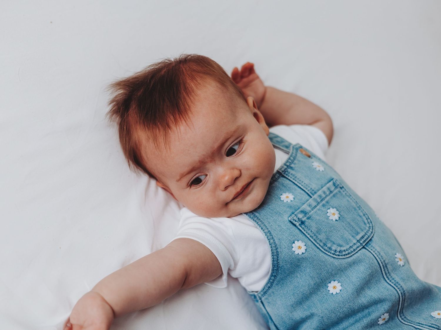 Adorable baby with red hair wearing a denim daisy overalls lying on a white background, smiling sweetly.