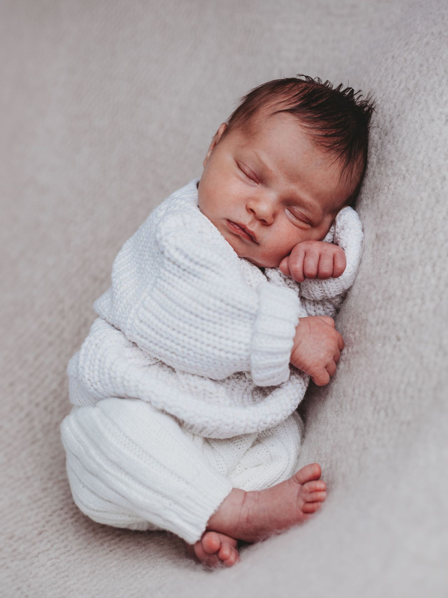 A peaceful sleeping newborn baby wearing a soft white knit sweater lies curled up on a textured gray blanket.