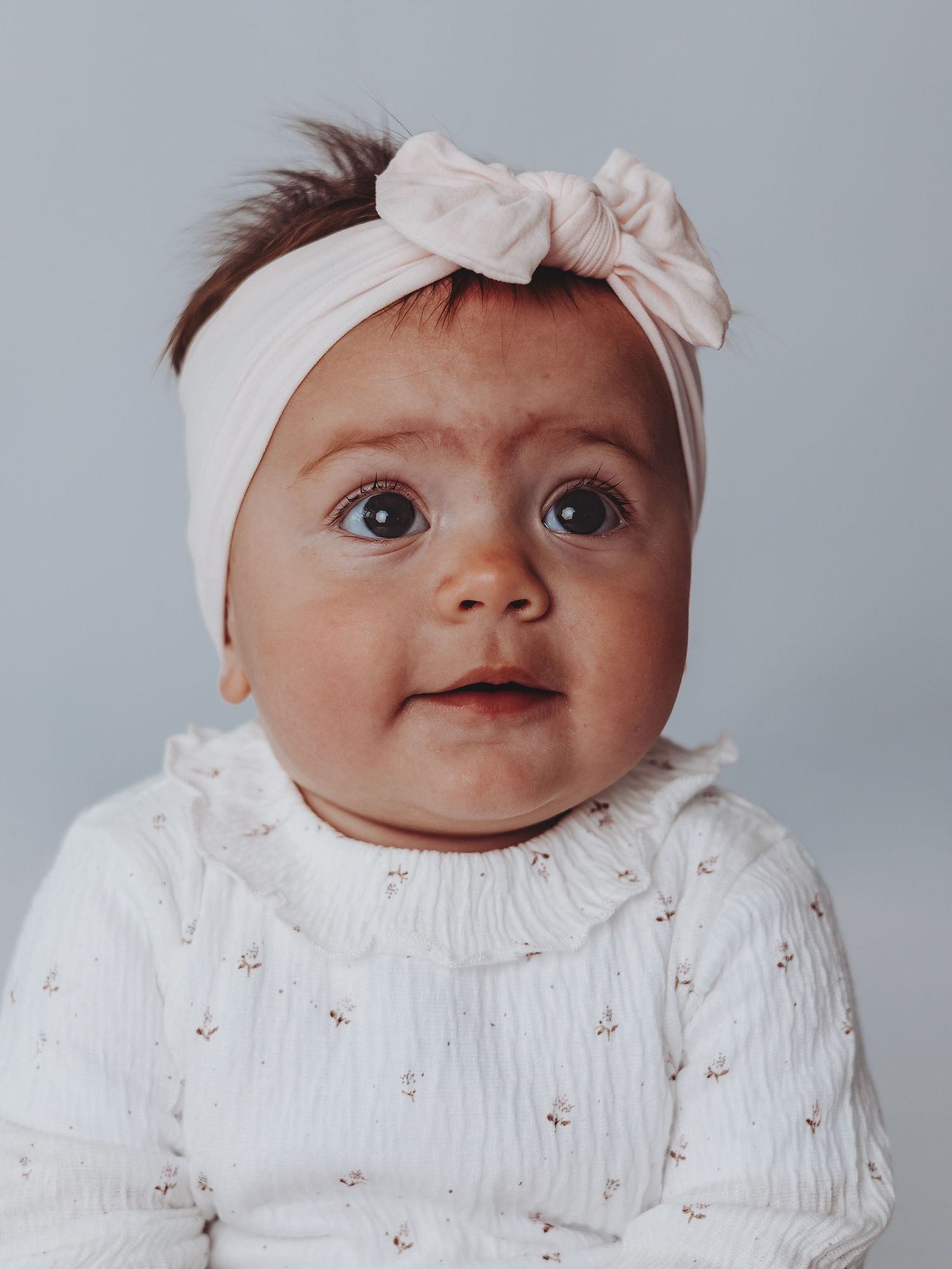 Baby wearing a white knit sweater and cream bow headband looks up with wide-eyed expression.
