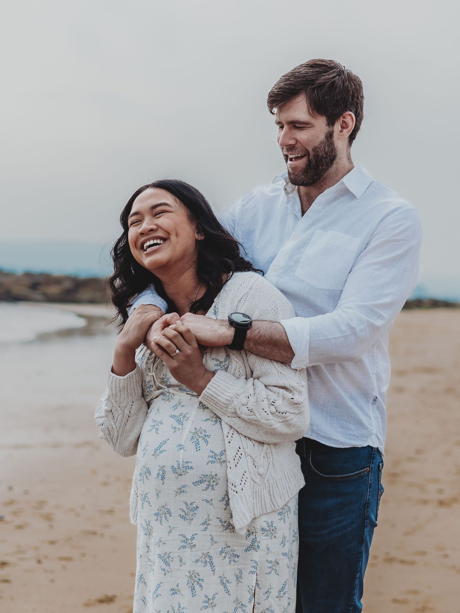 Romantic couple shares playful moments together on a cloudy beach in casual attire.