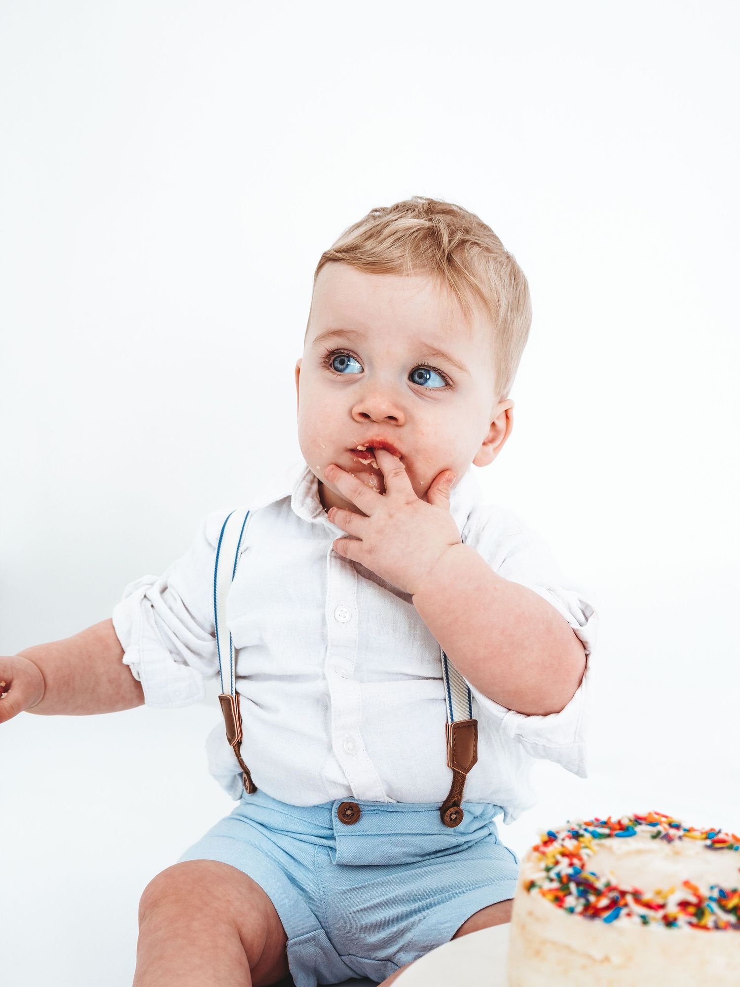 Adorable baby boy in white shirt and suspenders eating cake at his first birthday smash cake photo session.