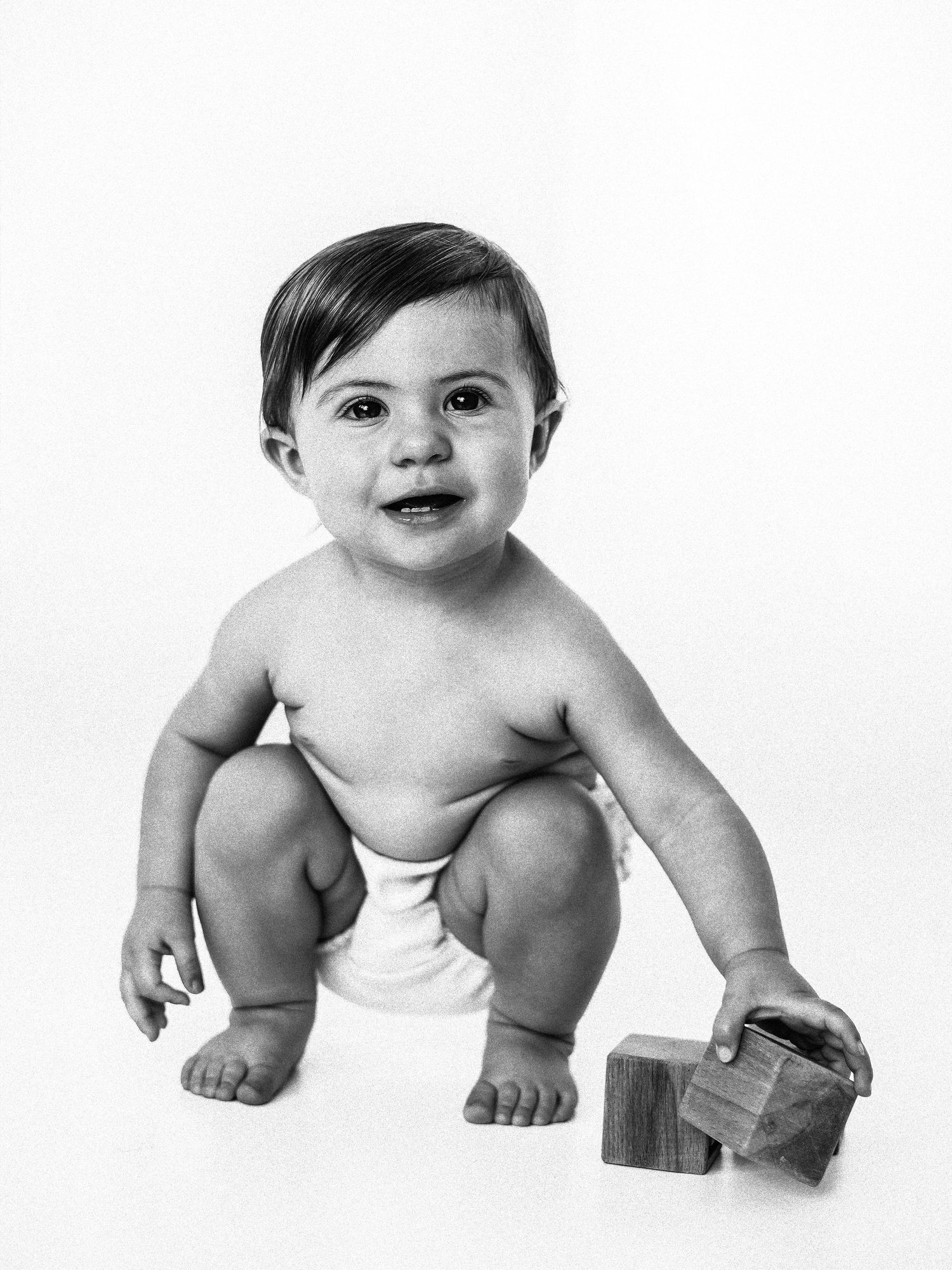 Black and white photo of a smiling baby in a diaper squatting and holding wooden blocks.