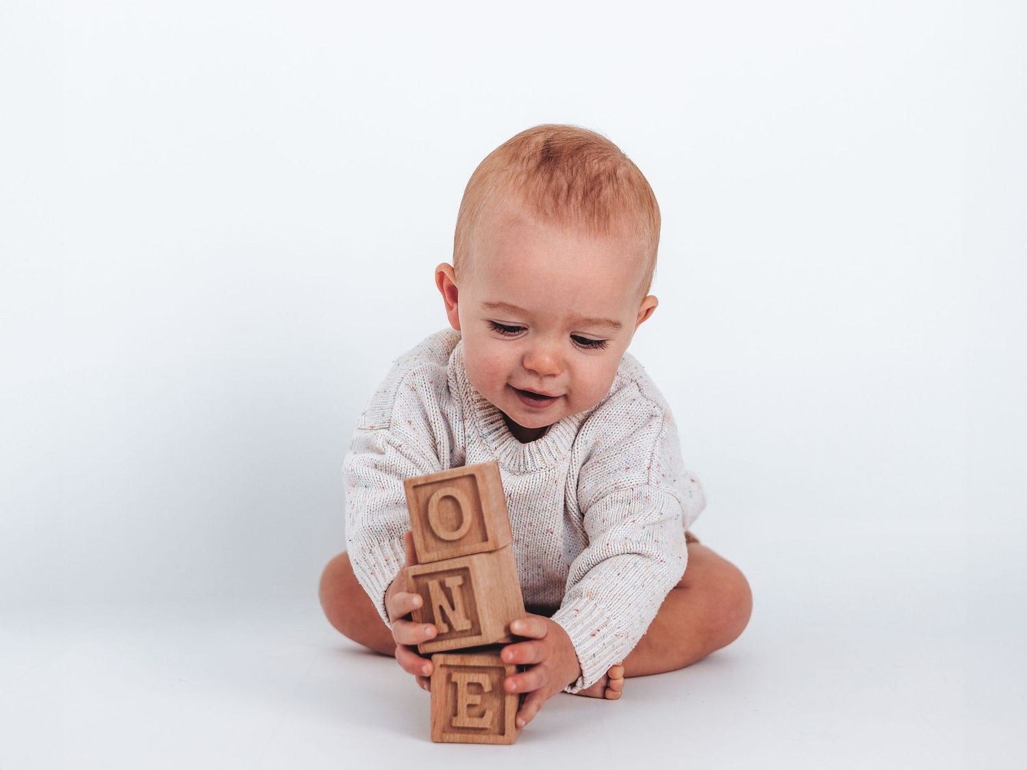 Baby in white sweater stacking wooden letter blocks spelling ONE, sitting on white background.