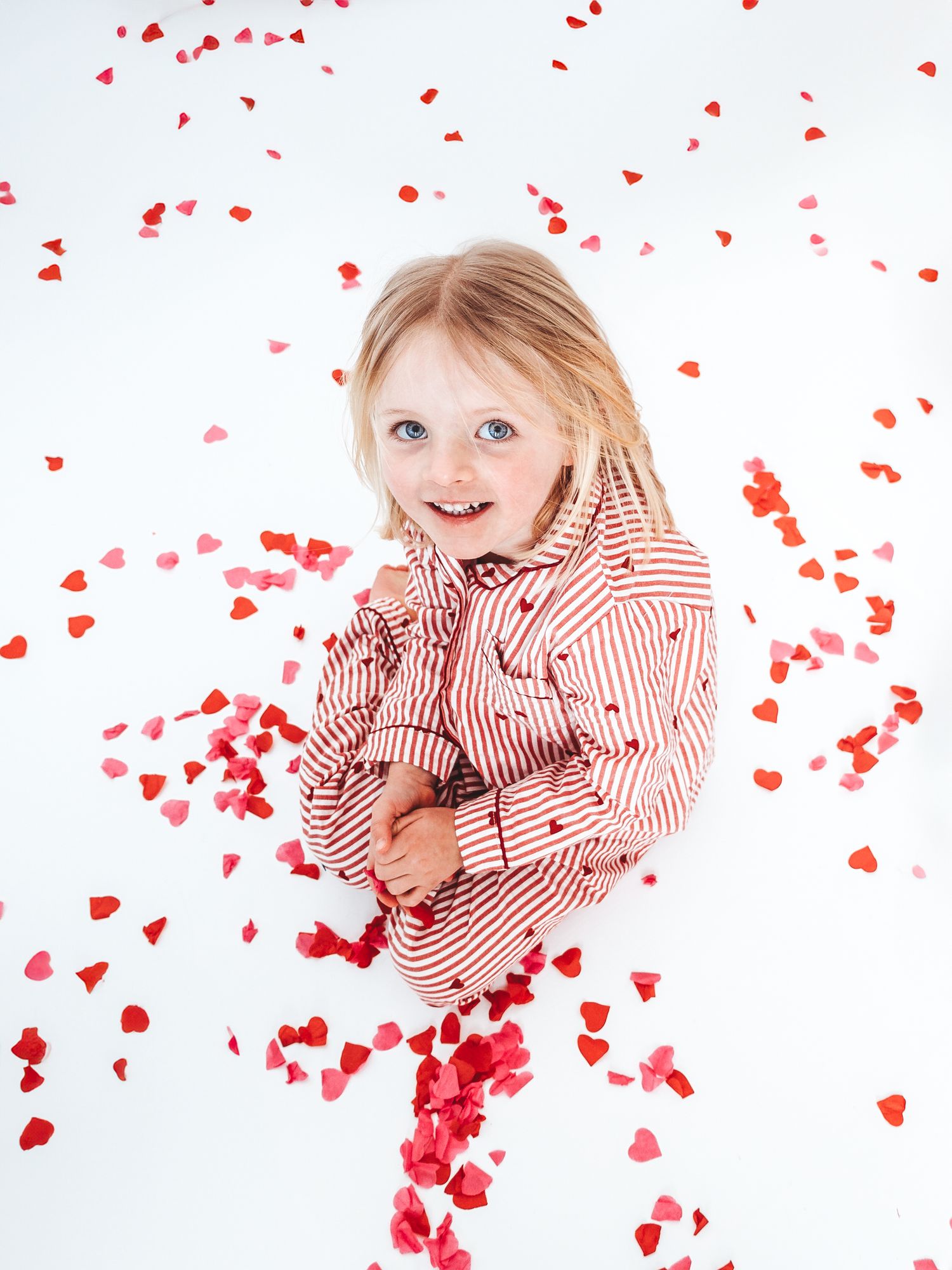 A cheerful young girl in a red striped shirt smiles surrounded by falling red heart-shaped confetti on white background.