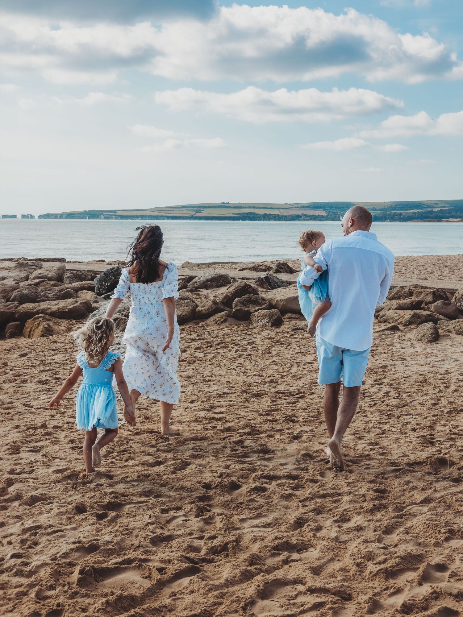 Family of four walking barefoot on sandy beach toward rocky shoreline, wearing light blue and white summer outfits.