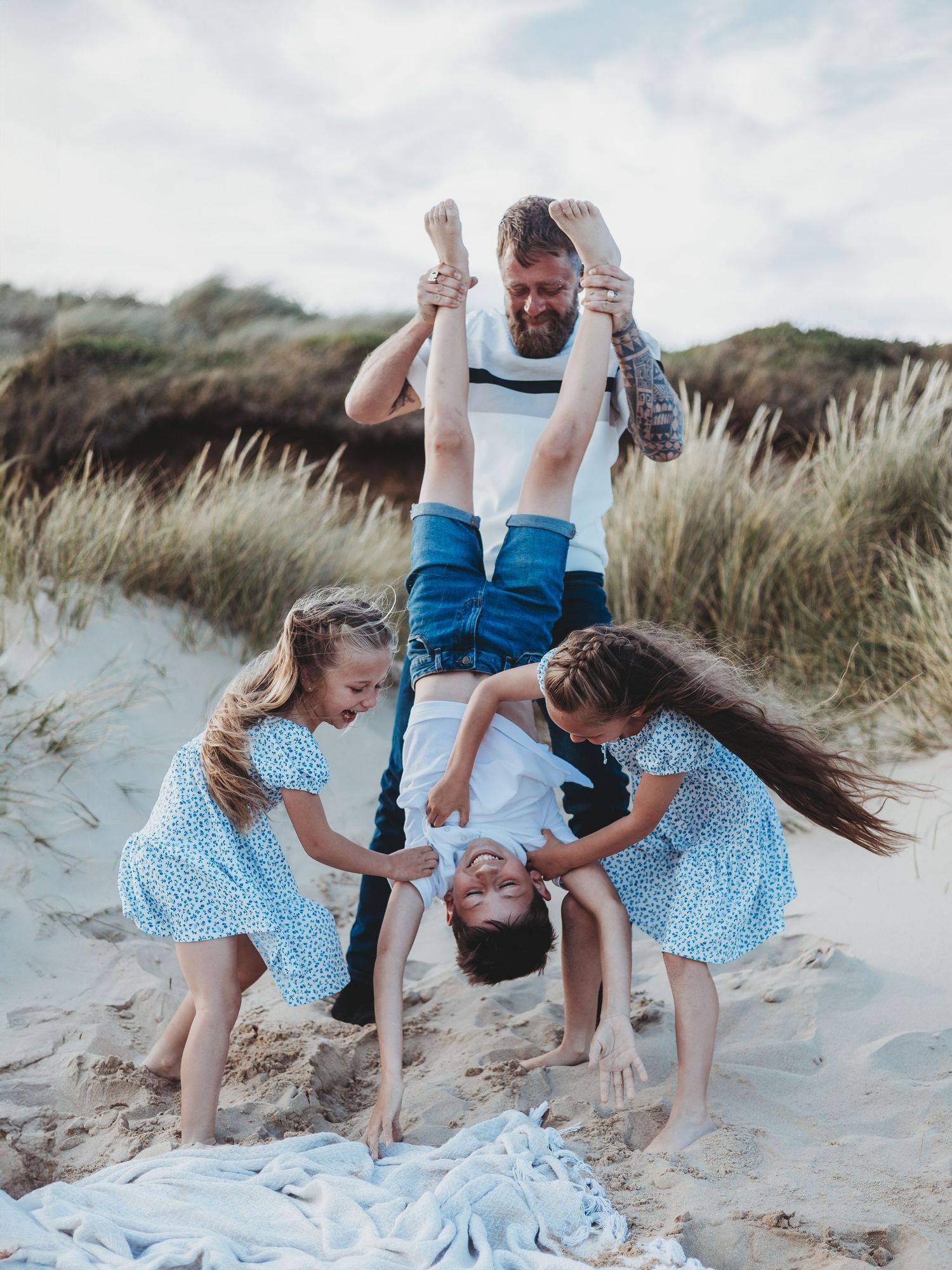 A bearded dad playfully holds a child upside down on a sandy beach while two girls in blue dresses laugh nearby.