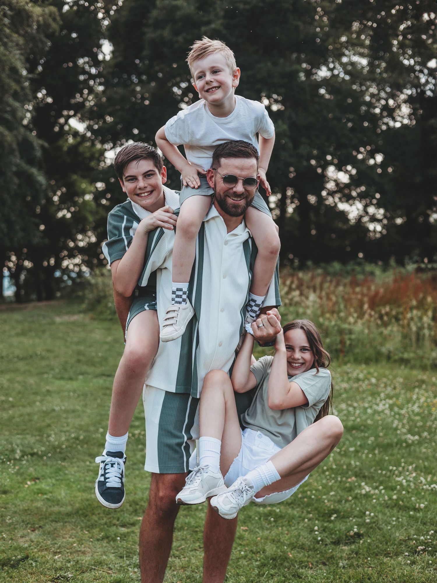 A man carries three children on his back and arms while posing in a lush green park setting.