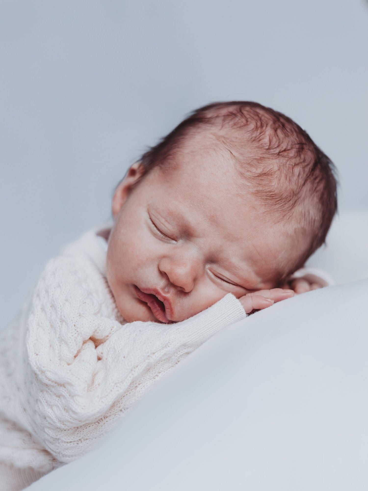A sleeping newborn baby wrapped in soft white fabric rests peacefully against a light background.