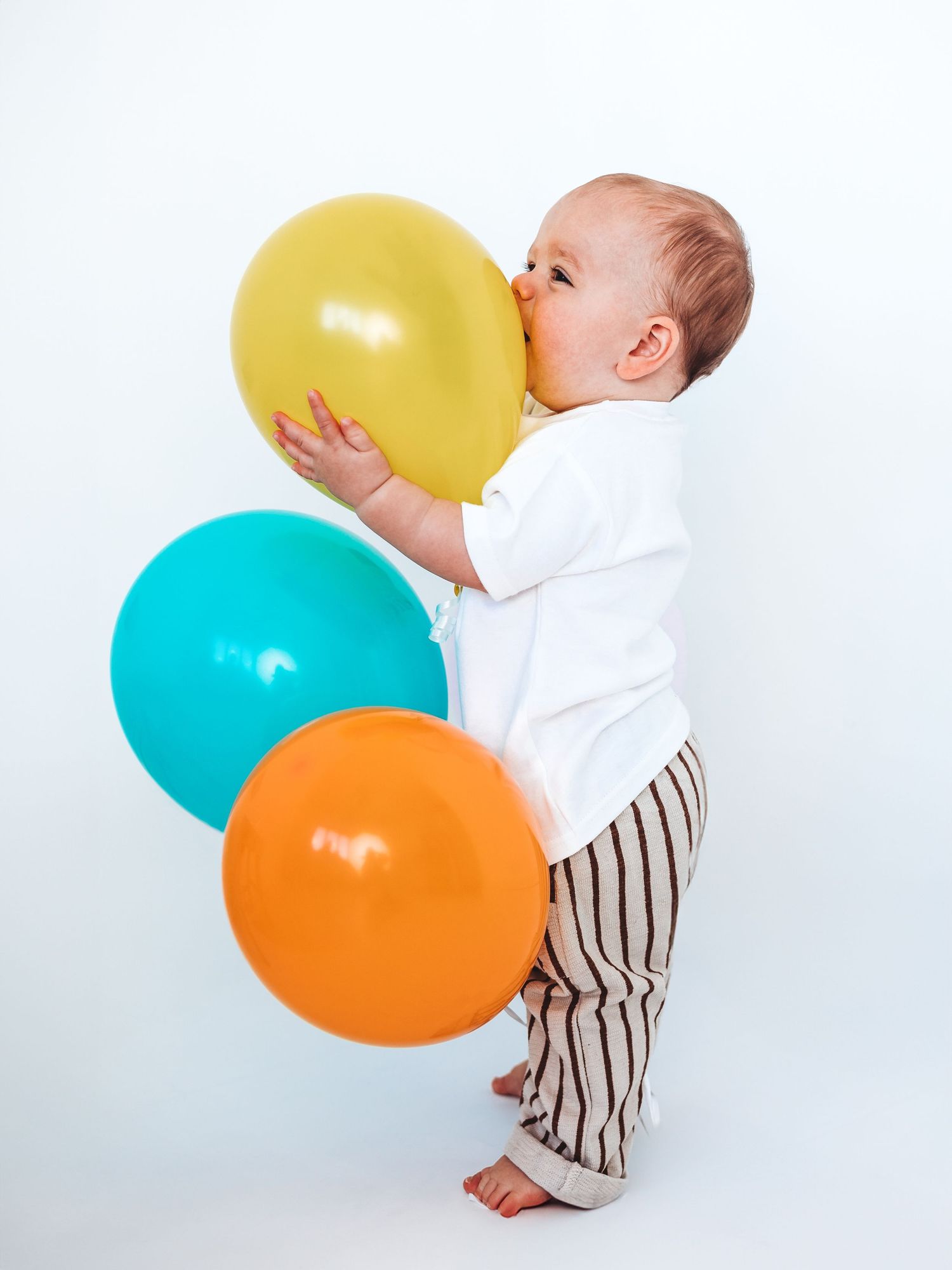 A cute baby in white onesie and striped pants playfully biting a yellow balloon with teal and orange balloons nearby.